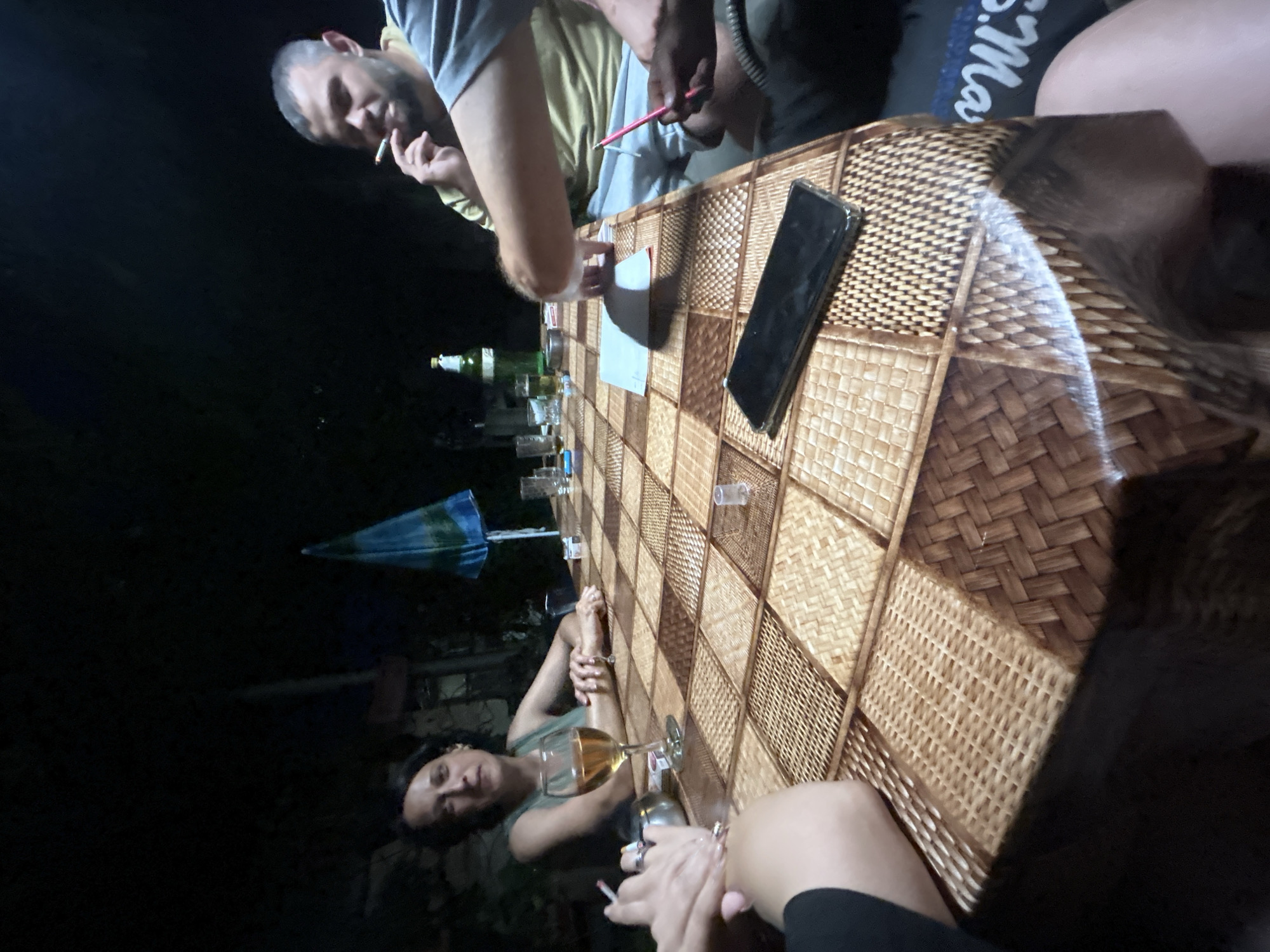 The image depicts a nighttime outdoor gathering around a table in Govedare, Bulgaria. Four individuals are visible, engaged in conversation and smoking. A woman in the foreground is holding a cigarette and looking towards the camera, while a man to her right is smoking and leaning into the table. The table itself is covered with a woven, checkered pattern and laden with items including a wine glass, bottles of beer, a smartphone, and ashtrays. The background is dark, with hints of foliage and what appears to be a closed umbrella. The scene suggests a casual, social event taking place in a residential or garden setting.