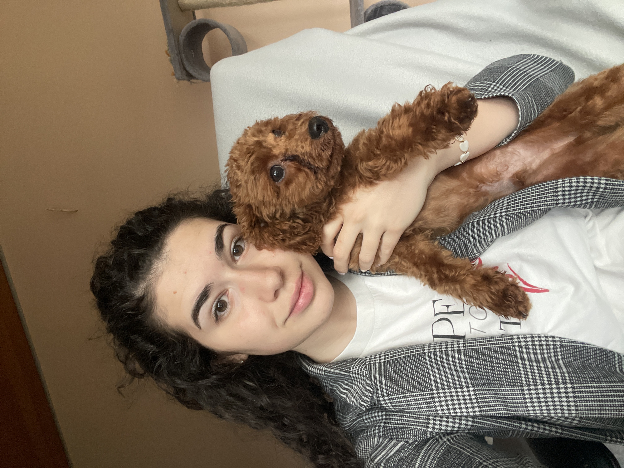 A young woman with dark curly hair lies down in an indoor setting, holding a small, reddish-brown poodle. She wears a white t-shirt with text, a grey-and-white plaid jacket, and a white bracelet. The background includes a light-colored wall and a cat tree. The location is impossible to determine, as there is no specific detail or landmark that is typical for the city of Sofia.