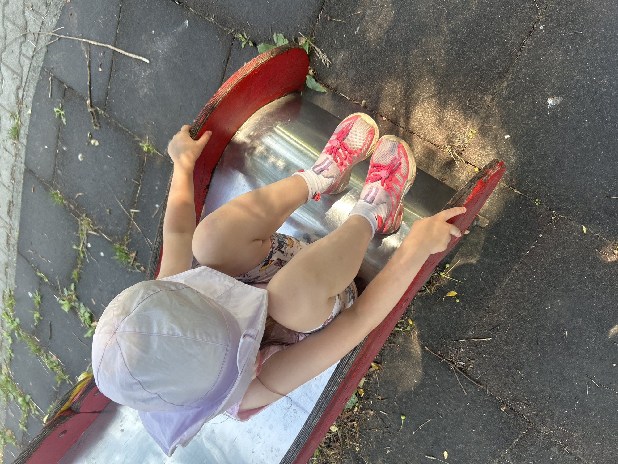 The image shows a young child wearing a light purple sun hat and pink and white sneakers sitting on a metal slide with red trim. The child is also wearing a pink top and patterned shorts. The slide is situated on a playground with a dark gray rubberized surface that is cracked and has some debris, including twigs and small green fruits, scattered on it. The background shows a paved area with some greenery. The scene is outdoors, likely in a public park or playground. The location in the city of Chaikovsky, Russia, is not discernible from the image itself.