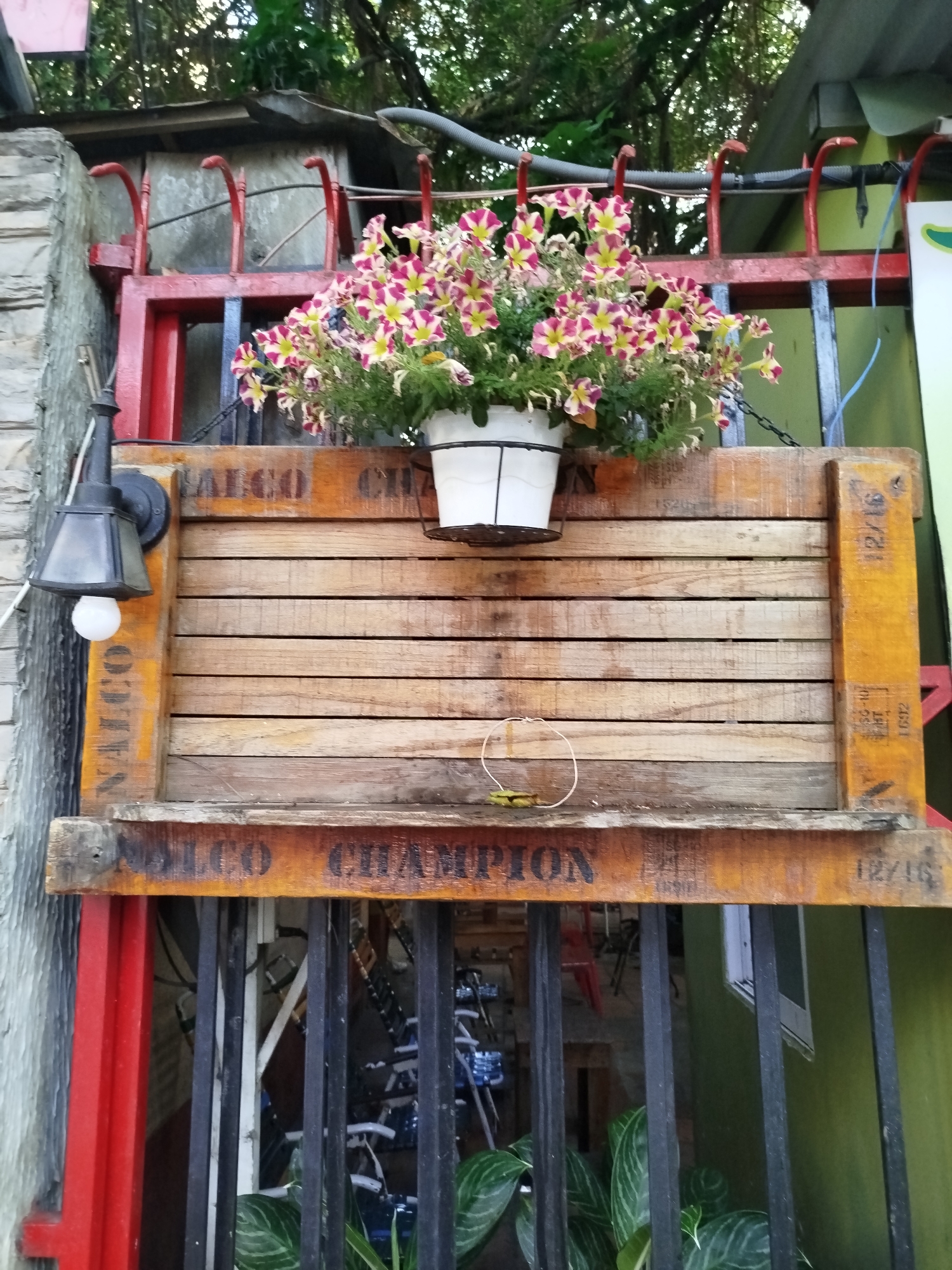 A low-angle shot captures a wooden signboard in front of what appears to be a gate, possibly in a residential area in Ho Chi Minh City, Vietnam. The gate features red and black bars. The signboard is rectangular with a plank design, having text "NALCO CHAMPION" stenciled on its upper and lower edges in a black font. A white flower pot filled with pink and yellow flowers hangs from the top of the signboard by chains. Attached to the left side of the board is a black industrial-style lamp with a white light bulb. A wire or hoop rests on the middle plank of the signboard, and some foliage is visible behind the gate. The signboard and the gate are surrounded by buildings and trees in the background.