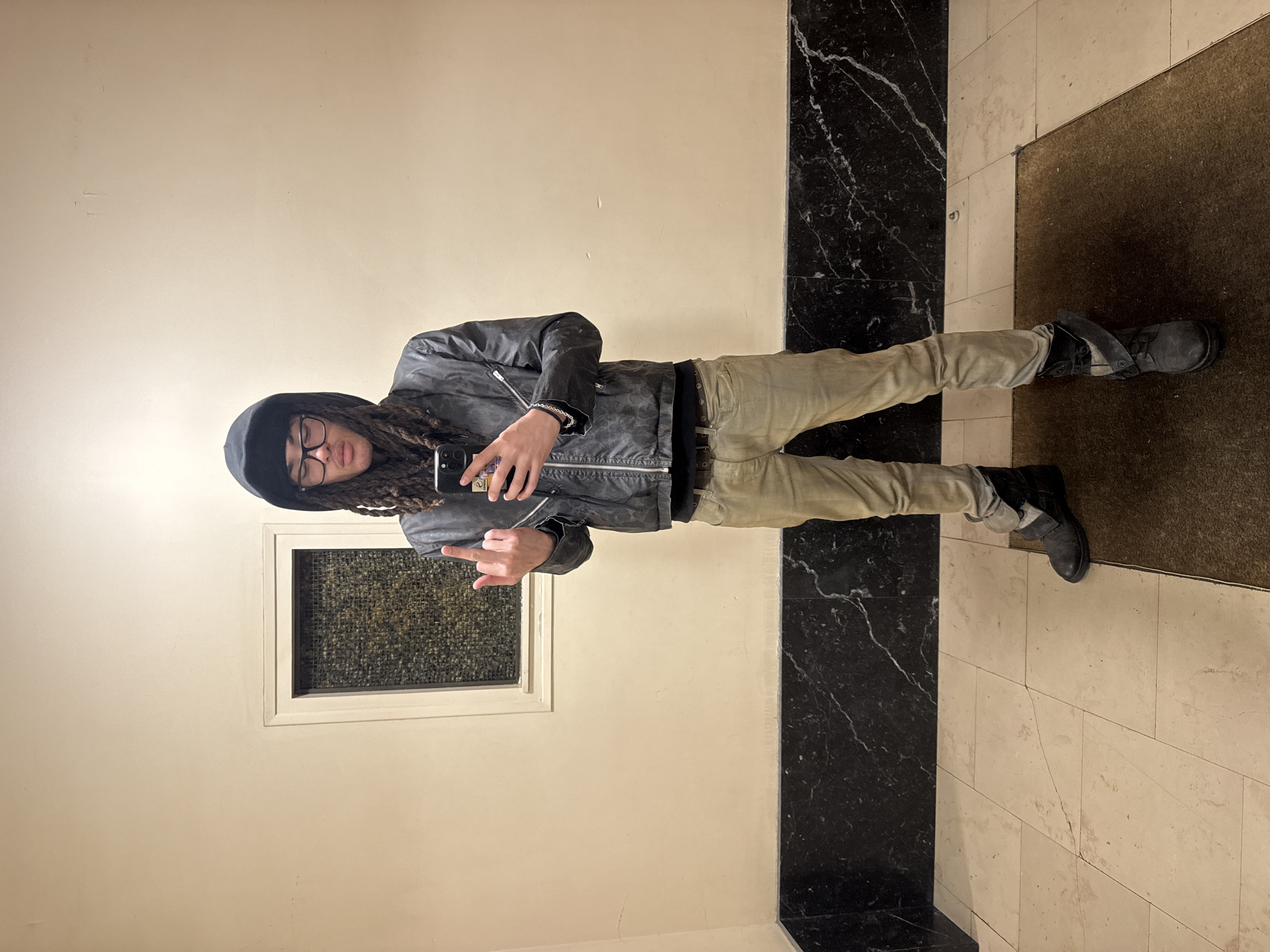 An individual with dark dreadlocks, wearing a black hooded top, black-rimmed glasses, a dark textured zippered jacket, light-colored pants, and dark boots, stands facing the viewer in an interior building space. The individual holds a dark-colored smartphone in their left hand, while their right hand is raised with the middle finger extended. The background consists of a light-colored wall above a dark, polished marble wainscoting with white veining. A recessed rectangular window with a dark mesh or textured glass insert is visible in the upper wall section to the left. The floor is covered with light-colored square tiles, and a dark brown floor mat occupies the lower right portion of the frame. The location is an interior hallway or common area of a building; no specific geographical identifiers for Paris, France, are visually present in the image.
