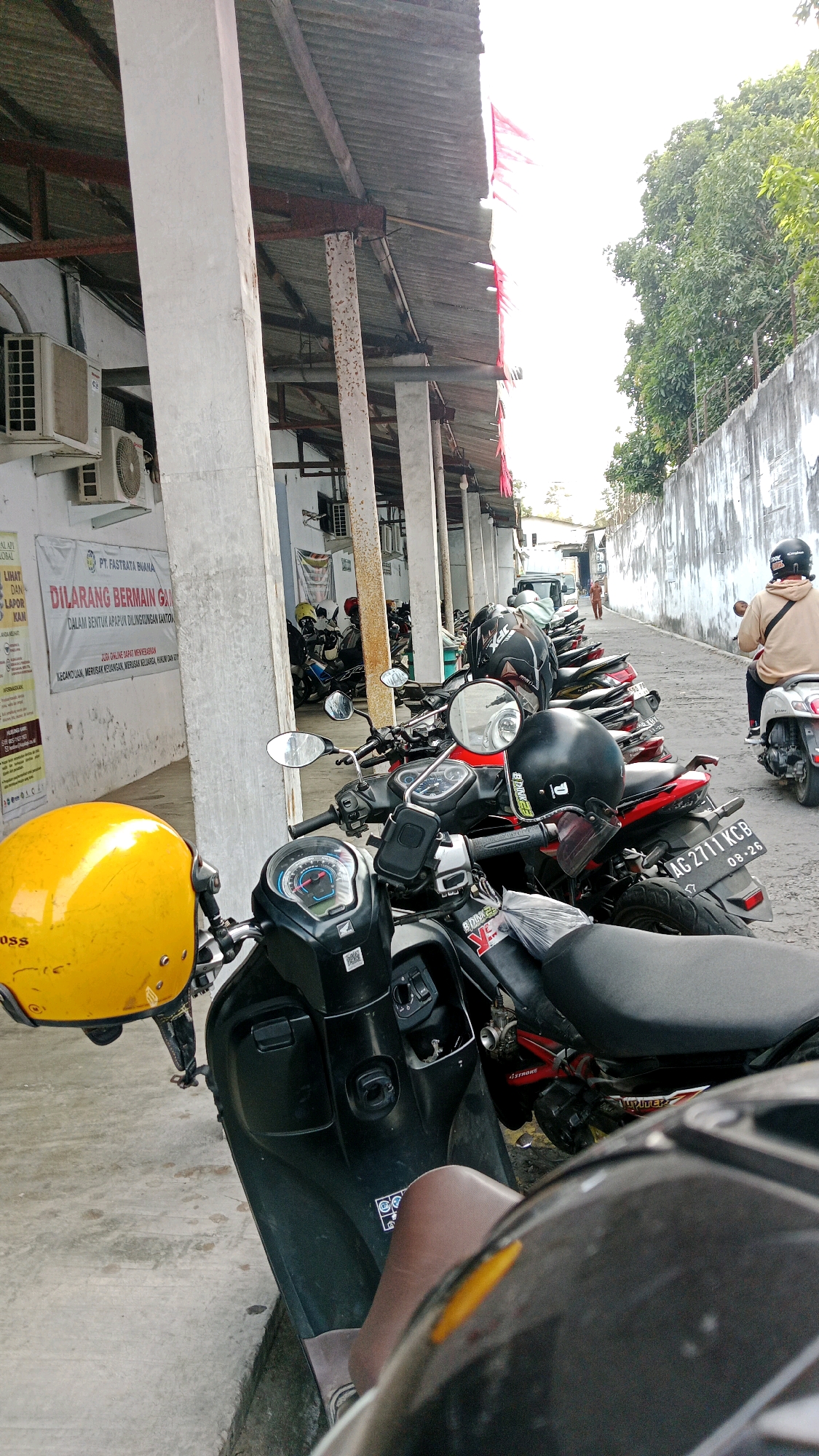 The image depicts an outdoor scene in a city, likely Blitar, Indonesia, characterized by a row of parked motorcycles under a covered walkway. The walkway is supported by concrete pillars and has a corrugated metal roof. The ground is paved with concrete.
In the foreground, a yellow motorcycle helmet rests on the handlebars of a black scooter. Several other motorcycles are parked in a line, some with helmets on their handlebars. One person is visible riding a white scooter in the background, wearing a tan jacket and a black helmet. Another person, wearing a light-colored shirt and dark pants, is walking further down the street.
A white wall runs along the right side of the image, with some greenery from trees visible above it. A sign with Indonesian text is affixed to the wall on the left side of the frame, near an air conditioning unit. The overall impression is that of a parking area adjacent to a building, likely in an urban environment.