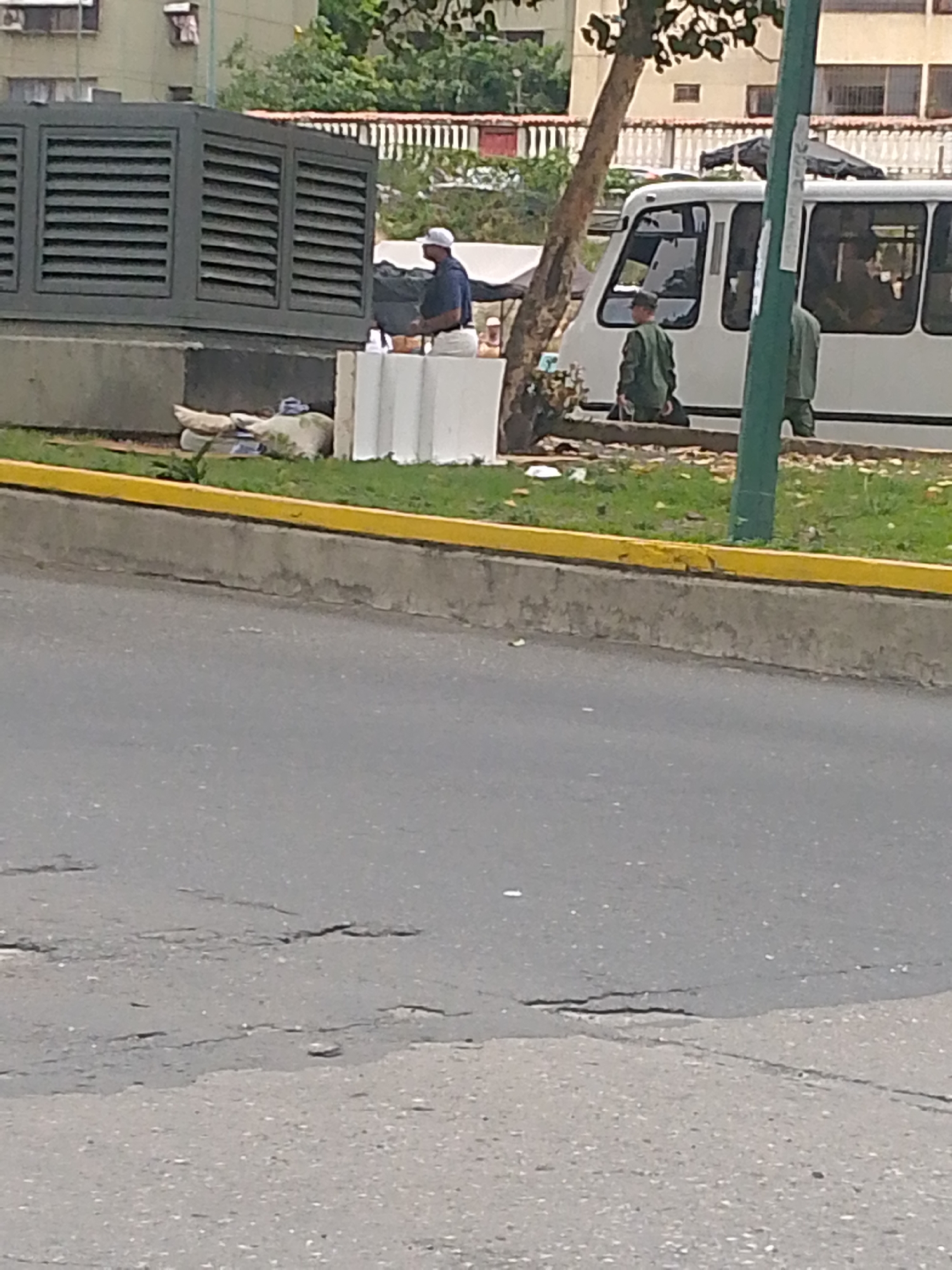 The image shows a street scene in Caracas, Venezuela. A white bus is parked on the side of the road, near a concrete median with a yellow curb. Two people dressed in military uniforms are standing near the bus. Another man in a white baseball cap and dark shirt is sitting on a concrete barrier next to a large grey structure with horizontal slats. Debris, including what appears to be bedding, is scattered around the base of the structure. The road surface is cracked and worn. A tree is positioned between the bus and the concrete barrier. The background features buildings and trees typical of an urban environment.