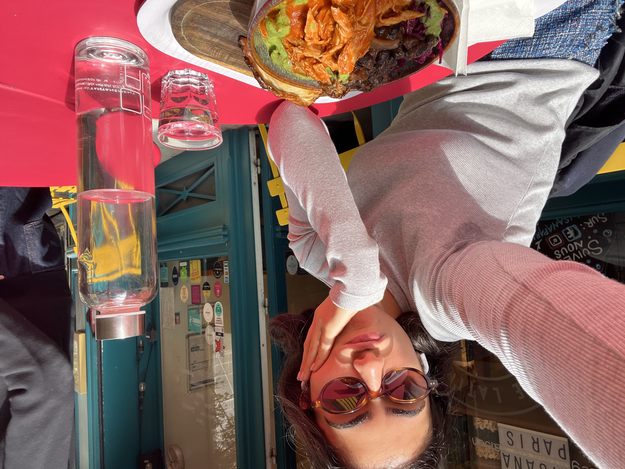 Here is a forensic description of the image:
The image is an upside-down selfie taken at a restaurant or cafe, likely in Paris, France, based on the signage visible in the lower right corner. The subject is a woman wearing sunglasses and a light gray long-sleeved shirt. She is holding her hand to her forehead. A plate of food with various components, including greens, shredded orange food, and dark food, is visible. There are glasses of water and an empty glass on a red table. Background elements include a turquoise-colored building facade with stickers and a display of signage in the window. There's a yellow chair nearby as well. The image is brightly lit, suggesting it was taken outdoors or near a window with natural light.