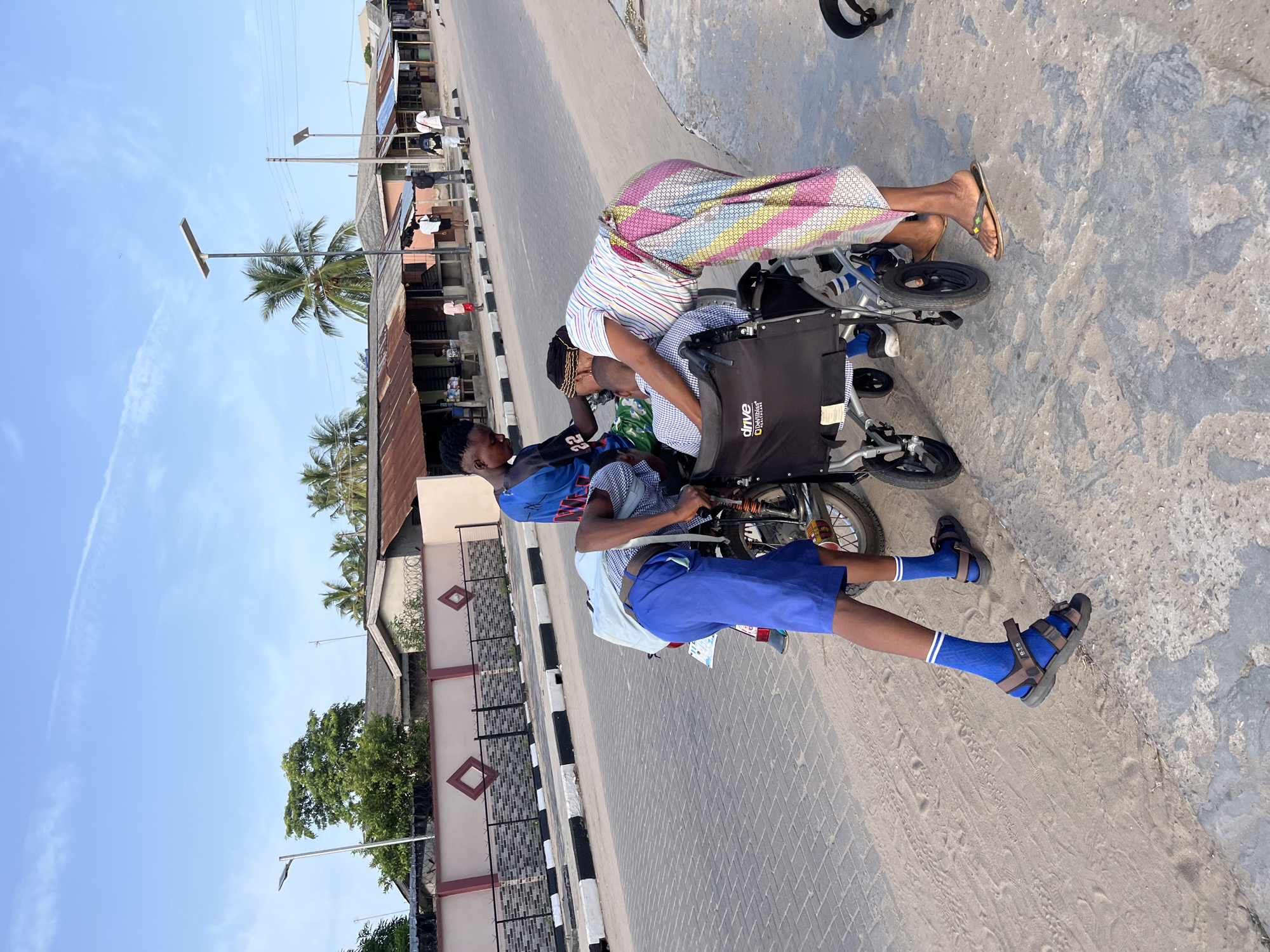 On a sunny day in Badagry, Nigeria, a young man in blue shorts and sandals pushes a wheelchair along a partially paved road. The wheelchair is occupied by an individual in a patterned garment, seemingly tending to someone in the chair. Other pedestrians can be seen further down the road. Adjacent buildings and palm trees suggest an urban setting. A black "Drive" logo is visible on the wheelchair's back. A motorcycle is visible behind the individual. A person wearing sandals is bent over the wheelchair.