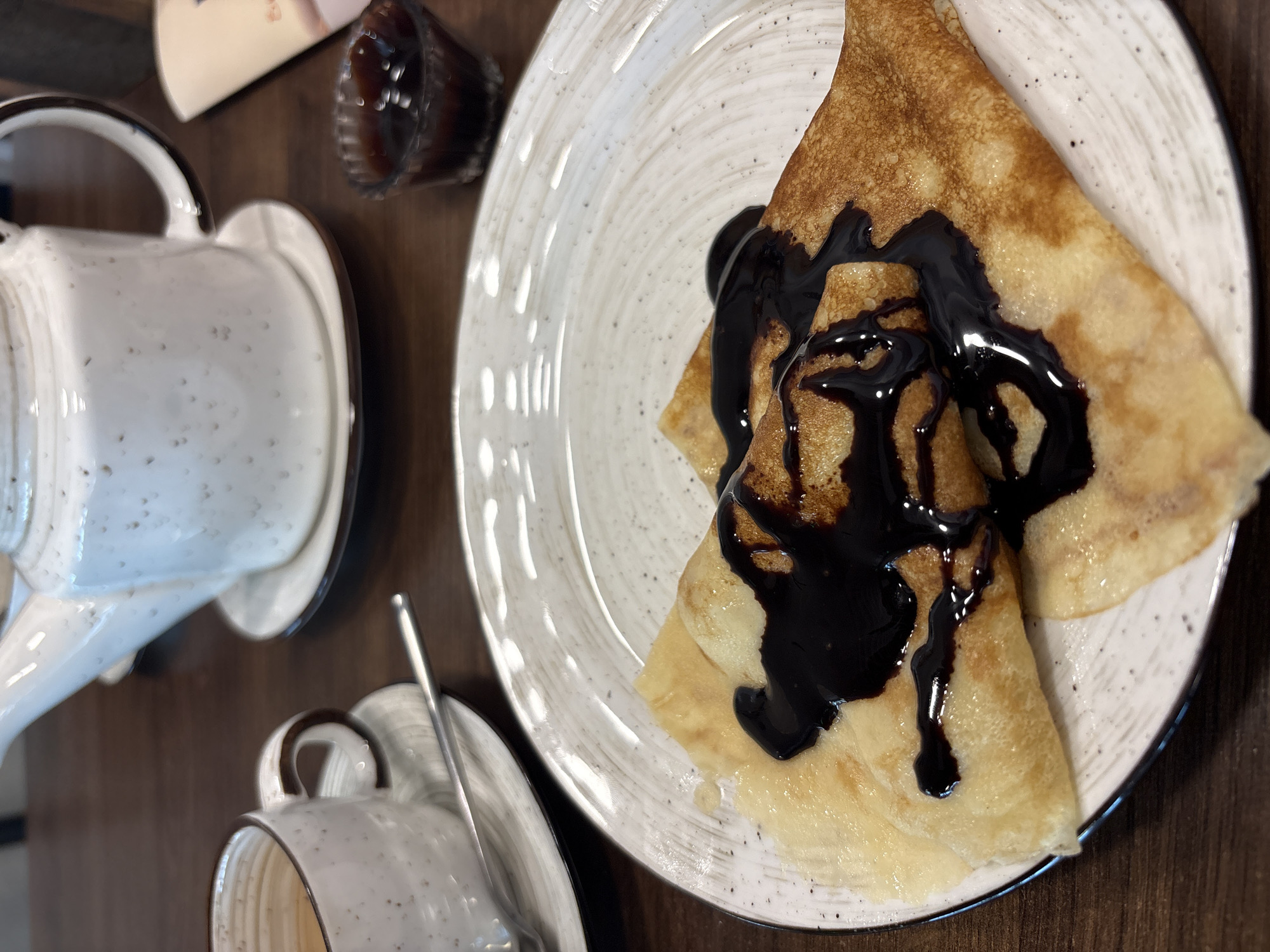 This image captures a close-up, elevated view of a breakfast or dessert setting on a dark brown wooden table in the city of Chaikovsky, Russia. In the foreground, two golden-brown pancakes, folded into triangular shapes, rest on a round, speckled white ceramic plate with a dark rim. A viscous, dark brown chocolate sauce is generously drizzled over both pancakes. To the upper left, a speckled white ceramic cup with a brown rim and handle sits on a matching saucer, containing a light brown liquid and a silver spoon. Further back and to the right, a matching speckled white ceramic teapot with a brown rim and handle is positioned on its own saucer. A small, dark, fluted glass containing a dark liquid, possibly jam or syrup, is visible on the far right. A blurred light-colored object is partially visible behind the small glass. No individuals are depicted in the scene.