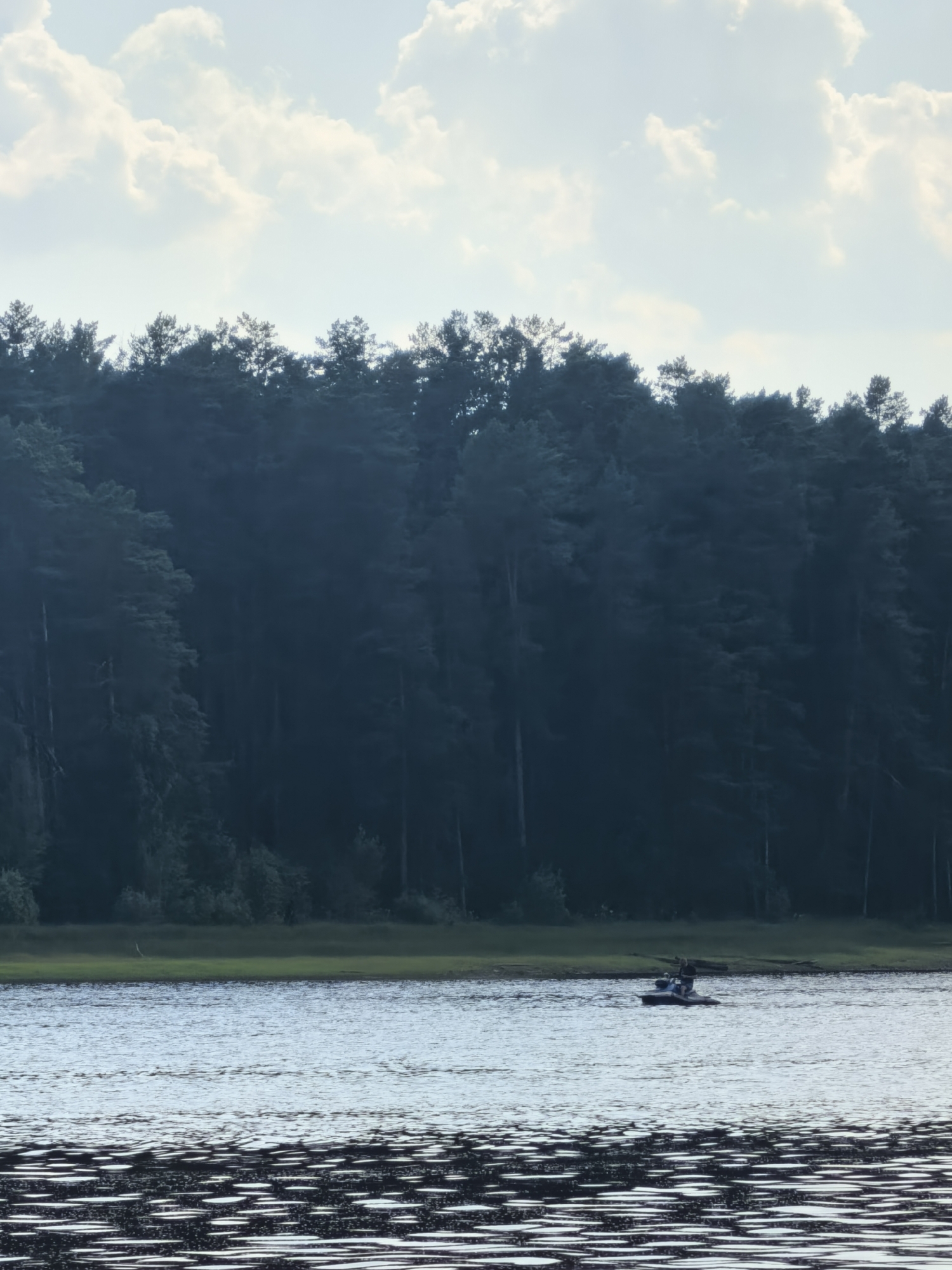 The image depicts a serene natural landscape near Sysert, Russia, characterized by a body of water in the foreground and a dense forest in the background. The water surface reflects the sky and surrounding environment, with ripples indicating gentle movement. In the middle distance, a small boat with two individuals is visible on the water, suggesting recreational activity. The forest, composed of tall, dark trees, creates a sense of depth and encloses the scene. The sky is partially obscured by scattered clouds, hinting at overcast conditions. The overall impression is one of tranquility and natural beauty.