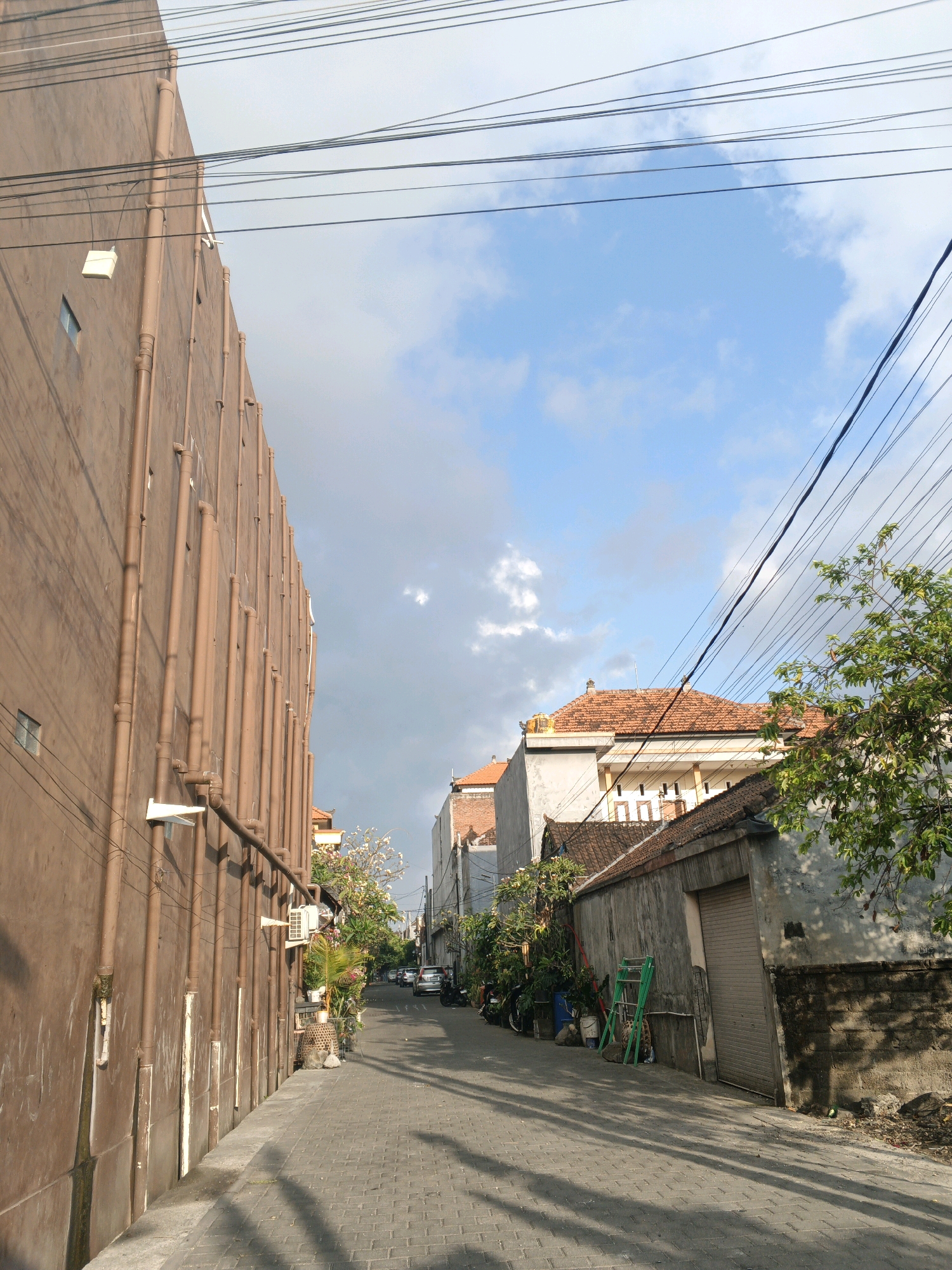An upward-angle, eye-level shot captures a narrow, paved alleyway in Denpasar, Indonesia, bordered by buildings and overhead utility wires. The left side of the alley is dominated by a tall, smooth, brown-walled building with numerous vertical brown pipes running down its exterior, spaced evenly. A white rectangular fixture is mounted near the top left of this building. The right side of the alley features a lower, weathered, grey-walled structure with a closed, ribbed metal roll-up door. Beyond this structure, other buildings with terracotta tile roofs are visible. Lush green trees and foliage are interspersed along the right side of the alley, with one prominent tree in the foreground on the right, its branches extending towards the center of the frame. A few parked motorcycles are visible further down the alley on the right, and a line of cars can be seen at a distance in the center. The ground is paved with rectangular grey bricks laid in a pattern. The sky above is blue with scattered white clouds. The lighting suggests it is daytime, with distinct shadows cast on the alleyway, primarily from the left-side building and trees.
