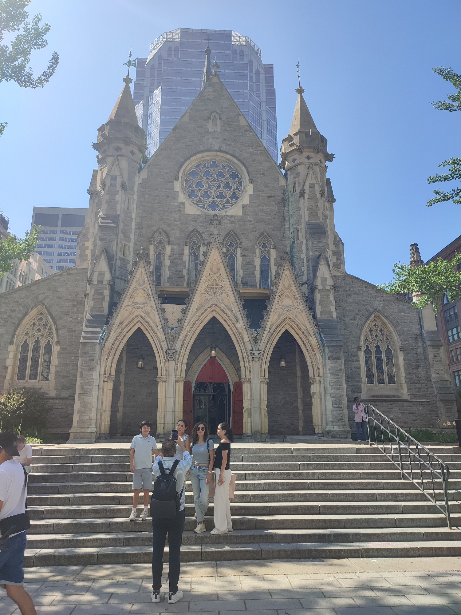 In Montreal, Canada, a group of people is captured on the steps of a large, stone church. The church features Gothic architectural elements, including pointed arches, a rose window, and spires. A modern glass skyscraper looms behind the church, indicating an urban setting.
On the steps, five individuals are visible. In the foreground, facing away from the camera, a person with a backpack is taking a photograph of the group. The group of four people is positioned on the middle steps. From left to right, the first person is a young male wearing a light blue polo shirt and shorts. Next to him is a young female in a white top and jeans, holding a mobile phone. To her right is a woman in a grey top and jeans, also holding a phone. The last person on the right is a female wearing a black off-the-shoulder top and white pants. To the right of the steps, a person in a pink shirt is standing near a railing, possibly engaged in a phone conversation. Two other individuals are partially visible on the far left of the image, with one appearing to be seated. The scene is bathed in bright sunlight under a clear blue sky.