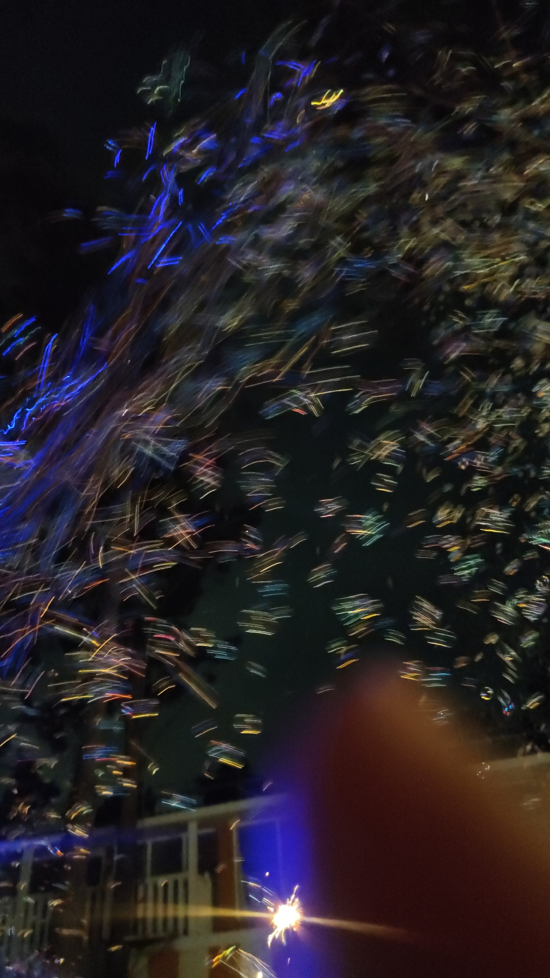 The image depicts a long exposure photograph taken at night in Beryozovka, Russia. Streaks of blue and yellow light suggest fireworks or a light show against a dark sky. In the lower portion of the image, a blurred building with white railings is visible, illuminated by blue light. A bright sparkler or small firework is also present, emitting light trails. A blurred reddish-brown object, likely a finger, is in the foreground, obscuring part of the scene. The overall impression is one of celebration or a festive event.