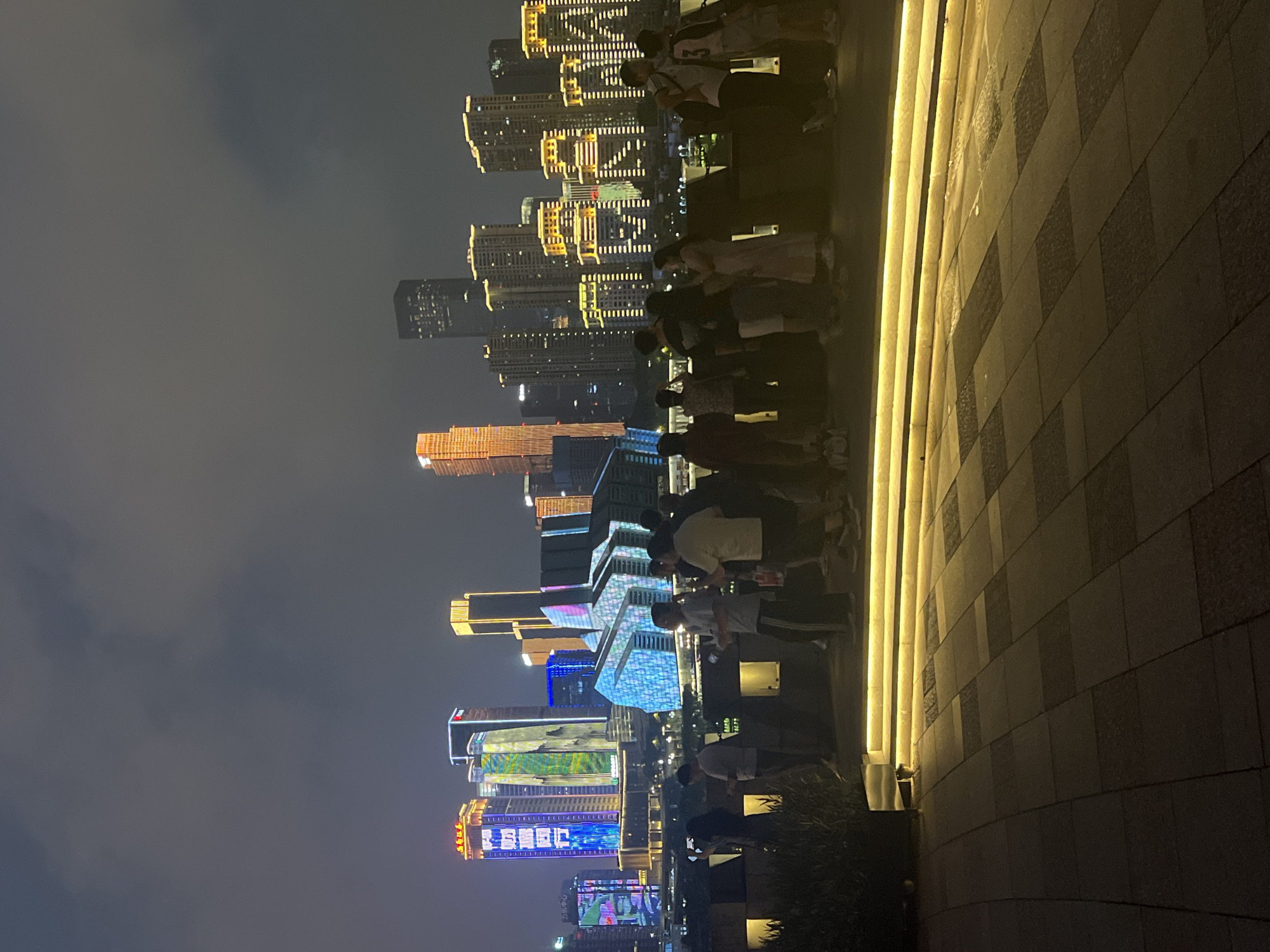 At night, a group of people stand on a stone-paved promenade overlooking a brightly lit cityscape. The buildings in the background are illuminated with colorful lights, some displaying patterns and text. The sky is dark and cloudy, creating a dramatic backdrop for the illuminated structures. The people are casually dressed and appear to be observing the view. The promenade has a raised section with integrated yellow lighting. The scene suggests a vibrant urban environment with a focus on nighttime attractions. The location is Yuzhong District, China.