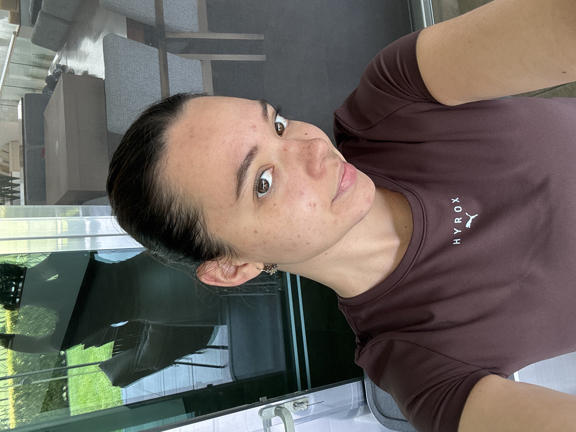 A selfie captures a young woman looking directly at the camera, with a slight, neutral expression. She has dark hair, styled away from her face, and brown eyes. Her skin appears fair with visible freckles and minor blemishes across her forehead, nose, and cheeks. She is wearing a dark brown, short-sleeved, crew-neck t-shirt. On the front of her shirt, the word "HYROX" is printed in white capital letters, accompanied by a white stylized logo resembling a leaping animal or figure. A small, decorative earring, possibly silver, is visible in her left ear.
The background consists primarily of a large glass panel, likely a window or sliding door, which creates reflections. Inside the reflection, modern furniture can be seen, including several light grey upholstered chairs with dark frames, and a light-colored rectangular object, possibly a table or storage unit. Beyond the glass, and also visible in reflection, is a bright green grassy area and some darker foliage, indicating an outdoor garden or lawn. The natural lighting suggests it is daytime. The image appears to be taken indoors, looking out. The provided location context is San Bernardino Tlaxcalancingo, Mexico.