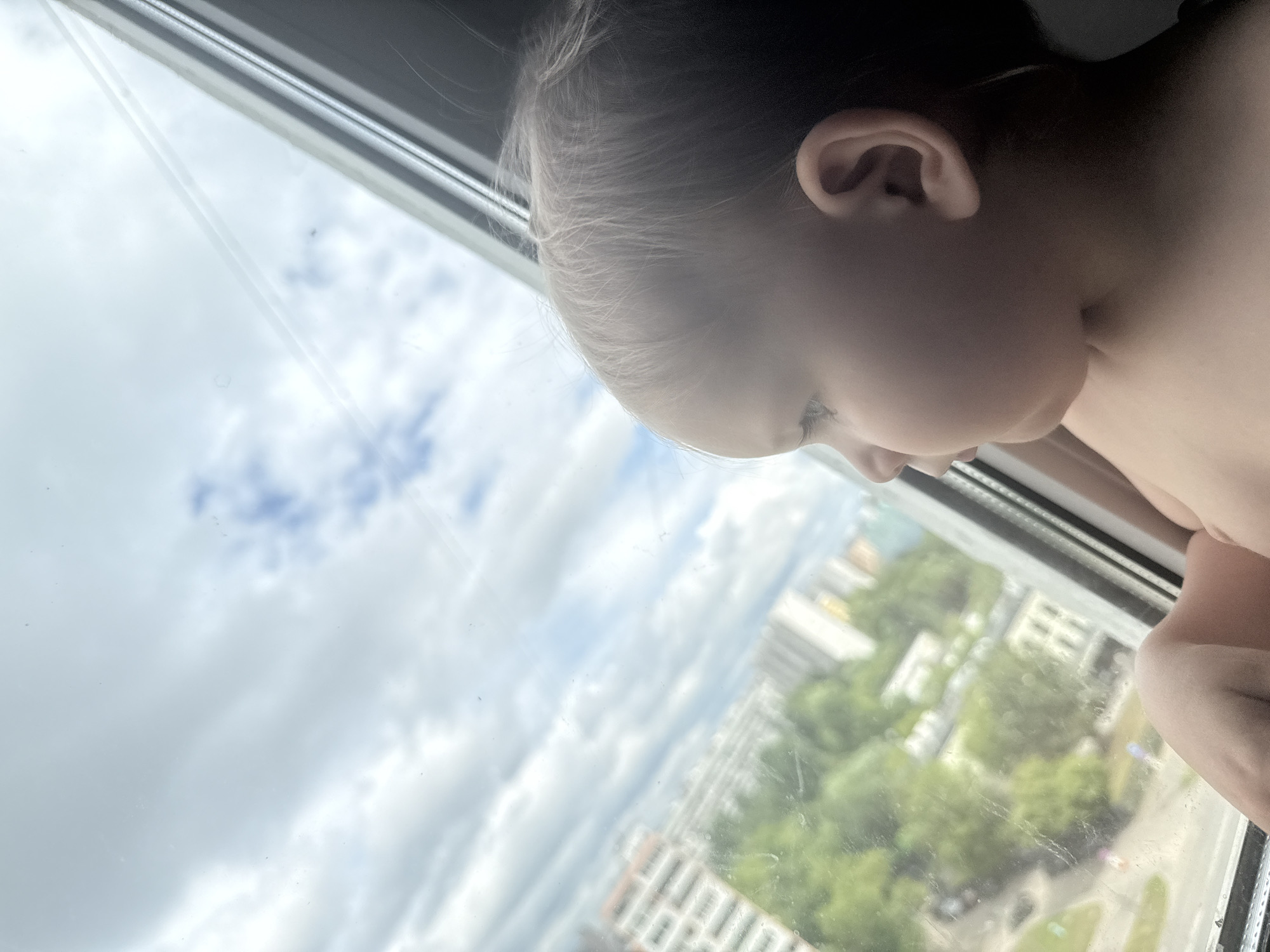 A close-up profile view of a young child looking out a window. The child's face is turned to the left, with their ear and the side of their head visible. The child has light-colored hair and appears to be wearing no clothing from the chest up. Outside the window, a blurry urban landscape is visible, featuring apartment buildings, trees, and a cloudy sky. The scene suggests a residential area, possibly a high-rise apartment building given the vantage point. The location is not specified as Moscow, Russia, or any other specific city.
