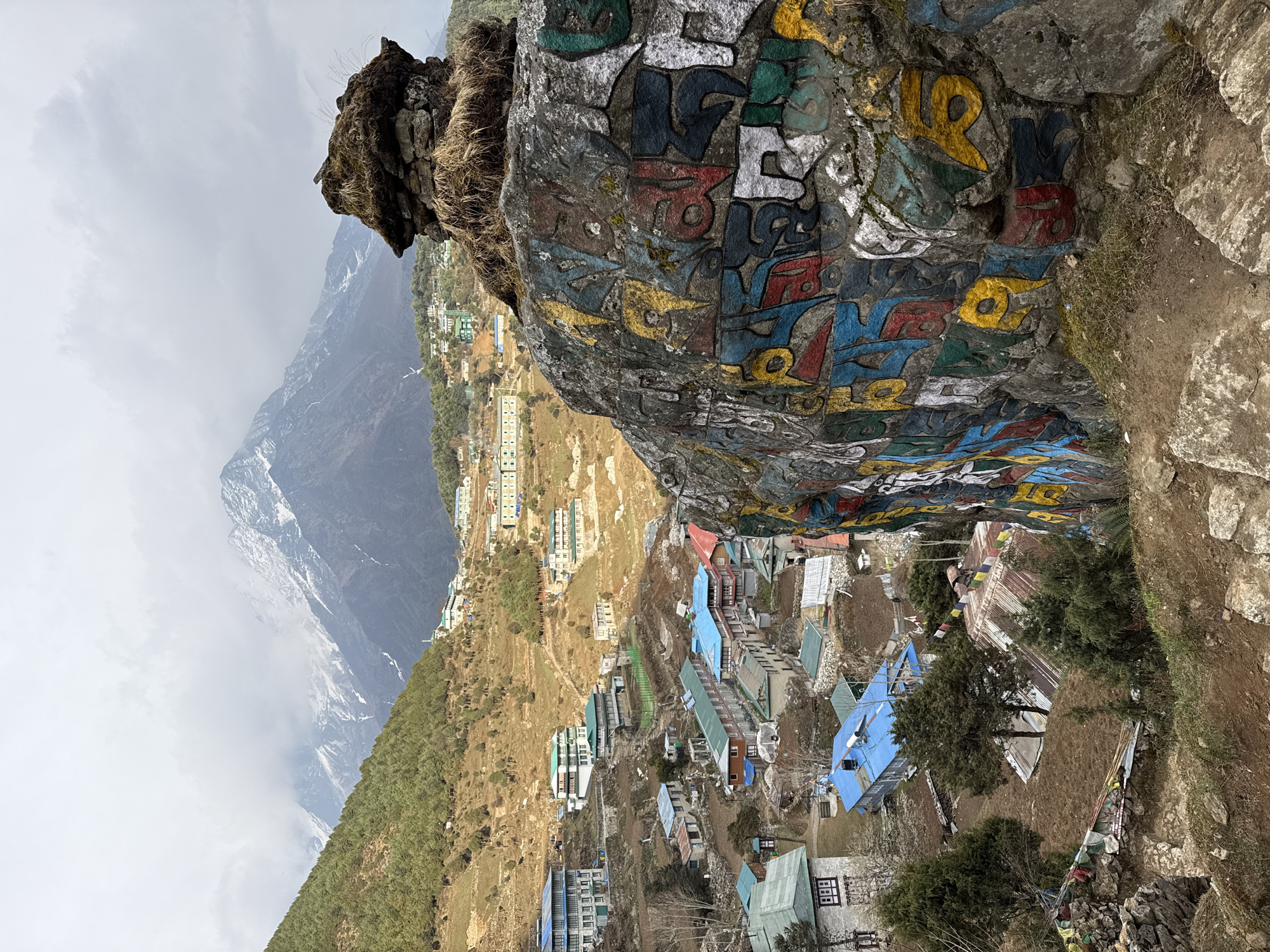 This image, presented with a vertical orientation (rotated 90 degrees counter-clockwise), captures a vibrant mountain village scene in Namche, Nepal, from an elevated perspective.
In the immediate foreground, a substantial rock face dominates the right side of the frame. This natural rock is extensively painted with colorful Tibetan script, predominantly in red, yellow, blue, white, and green hues. The script appears to be a religious mantra, characteristic of Mani stones found in Himalayan regions. The rock surface is rough and textured, with patches of moss and dry grass growing in crevices, particularly at its top edge.
Beyond the painted rock, the midground reveals the sprawling village of Namche Bazaar, nestled on a steep, terraced hillside. Numerous buildings, likely lodges, homes, and shops, are visible. They feature a variety of roof colors, including bright blue, red, green, and corrugated metal, indicating different construction materials and styles. Many structures are multistoried, and some prayer flags are discernible, fluttering among the buildings, especially near a cluster of trees. The village structures ascend the slope, with some buildings appearing more modern and others more traditional.
The background showcases the rugged Himalayan landscape. Towering, dark mountains rise majestically, with their peaks partially obscured by an overcast, cloudy sky. Snow patches are visible on the higher slopes, indicating a significant altitude and cool climate. Dense green forests cover the lower mountain flanks and parts of the hillsides surrounding the village. The sky is largely gray and diffused, suggesting an overcast day, potentially late morning or early afternoon, though no direct sun is evident.
No people or specific activities are visible within the frame, emphasizing the grand scale of the natural landscape and the architectural presence of the village. The scene evokes a sense of tranquility and the resilient human settlement in a dramatic mountain environment.