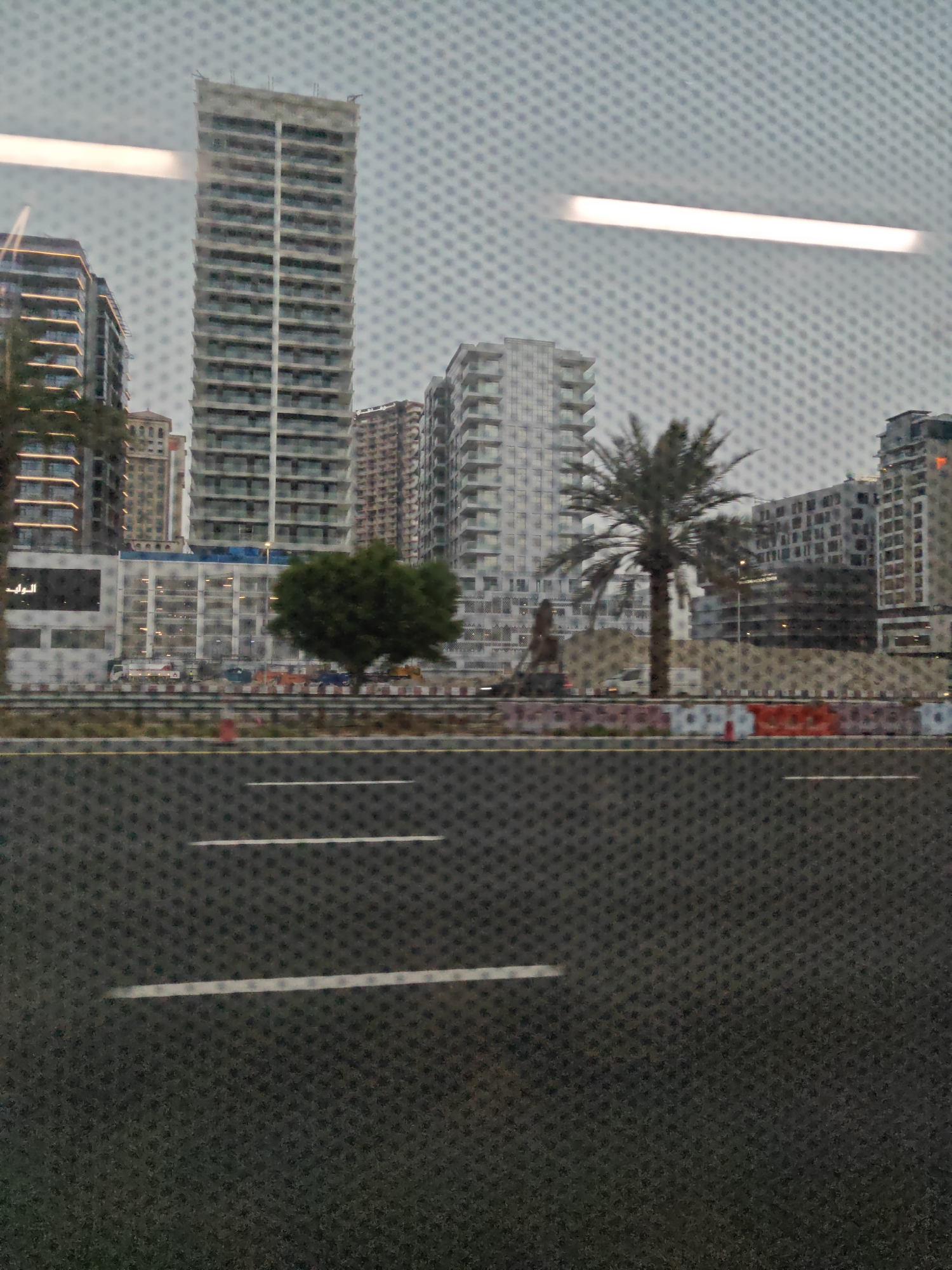 The image depicts a street-level view of a construction zone in Dubai, United Arab Emirates, as seen through a textured glass surface. In the background, a row of modern high-rise buildings stands against a hazy sky. One prominent building in the center appears to be under construction, with exposed concrete floors and unfinished facade elements. To its left and right are other residential or commercial towers with balconies and windows. A tall palm tree is visible between some of the buildings.
In the mid-ground, a road with white lane markings is present, separated from the buildings by a barricade. The barricade consists of orange and white plastic barriers and some construction fencing. A few vehicles, including what appear to be construction trucks and a white van, are visible behind the barricade. A single, round green tree stands near the center of the frame, adjacent to the road.
The foreground is dominated by the textured glass, which creates a grid-like pattern overlaying the entire scene. This glass appears to be a window, possibly of a vehicle, looking out onto the street. The overall impression is of a dynamic urban environment in a state of ongoing development. There are no discernible individuals in the image.
