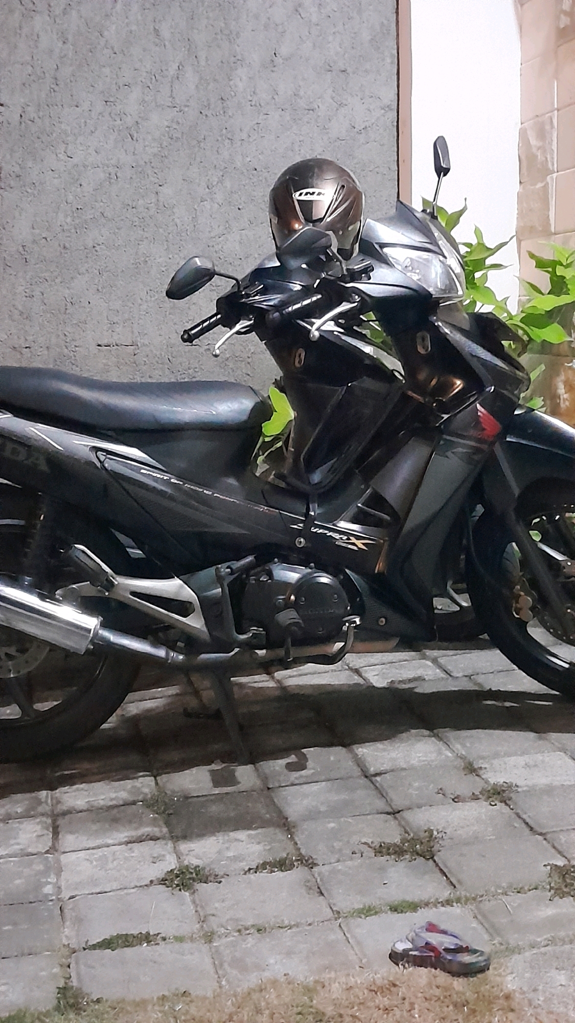 A side-view, medium shot captures a black Honda motorcycle parked on a patterned paved surface. A dark brown helmet with a metallic sheen rests on the handlebars. The motorcycle exhibits various details, including its seat, engine, exhaust pipe, and the "SUPRA X" emblem on its side. In the background, a textured gray wall and a white tiled structure can be seen, along with lush green foliage. The ground in the foreground is a mix of dry grass and the same patterned paving stones. A single flip-flop is visible on the ground in the lower right corner of the frame. The lighting suggests it may be dusk or an overcast day.