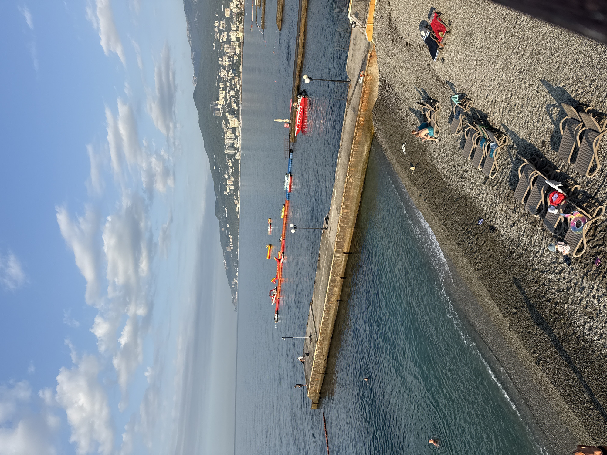 The image captures a sunny day at a pebble beach in Yalta, Ukraine. In the foreground, numerous empty lounge chairs are arranged on the beach, with a few people relaxing on them. The sea is calm, with gentle waves lapping the shore. A concrete pier extends into the water, where a few individuals are fishing or swimming. Further out in the sea, a colorful inflatable water park with various structures is visible, along with a red boat. In the background, the city of Yalta stretches along the coastline, with buildings nestled against a lush green mountainside. The sky is partly cloudy, with patches of blue.