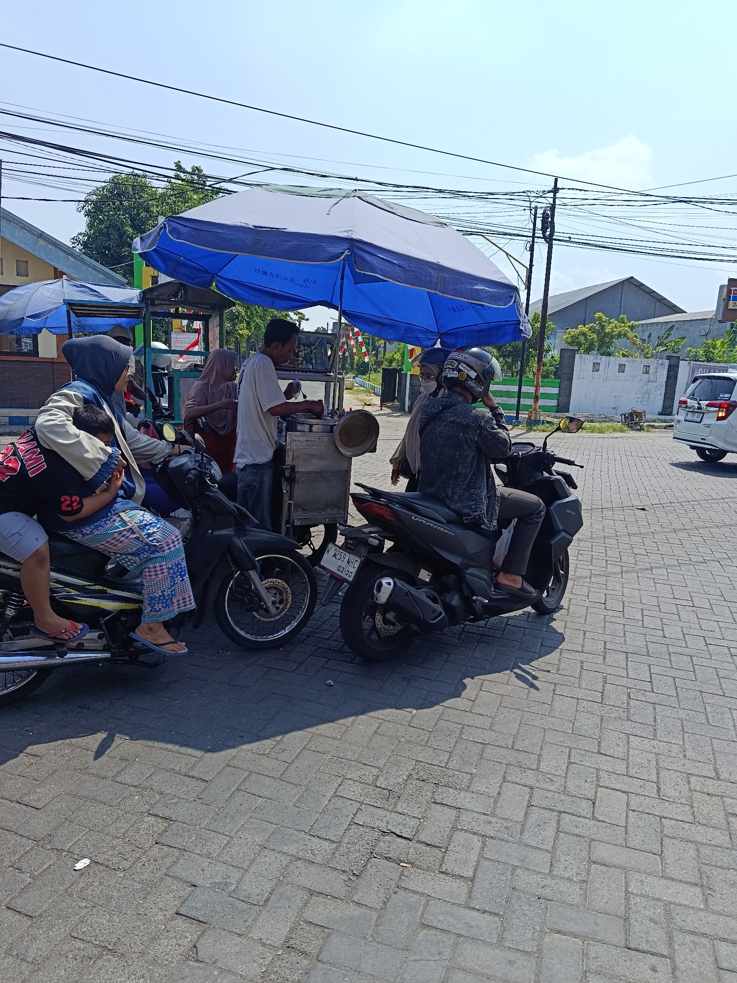In Keboansikep, Indonesia, on a sunny day, a food cart vendor is operating under a blue and white umbrella on a paved street. Several people are gathered around the cart. One person, wearing a blue hijab and a cream-colored jacket, is seated on a motorcycle with a child on their lap. The child is wearing a black shirt with the number "22" and patterned pants. Another person, a man in a white t-shirt and jeans, is standing at the food cart, seemingly serving food. A woman in a tan hijab is also near the cart. To the right, another person wearing a denim jacket and helmet is seated on a black motorcycle. In the background, buildings and utility poles with numerous wires are visible. A white car is parked further down the street. The ground is covered in interlocking brick pavers.
