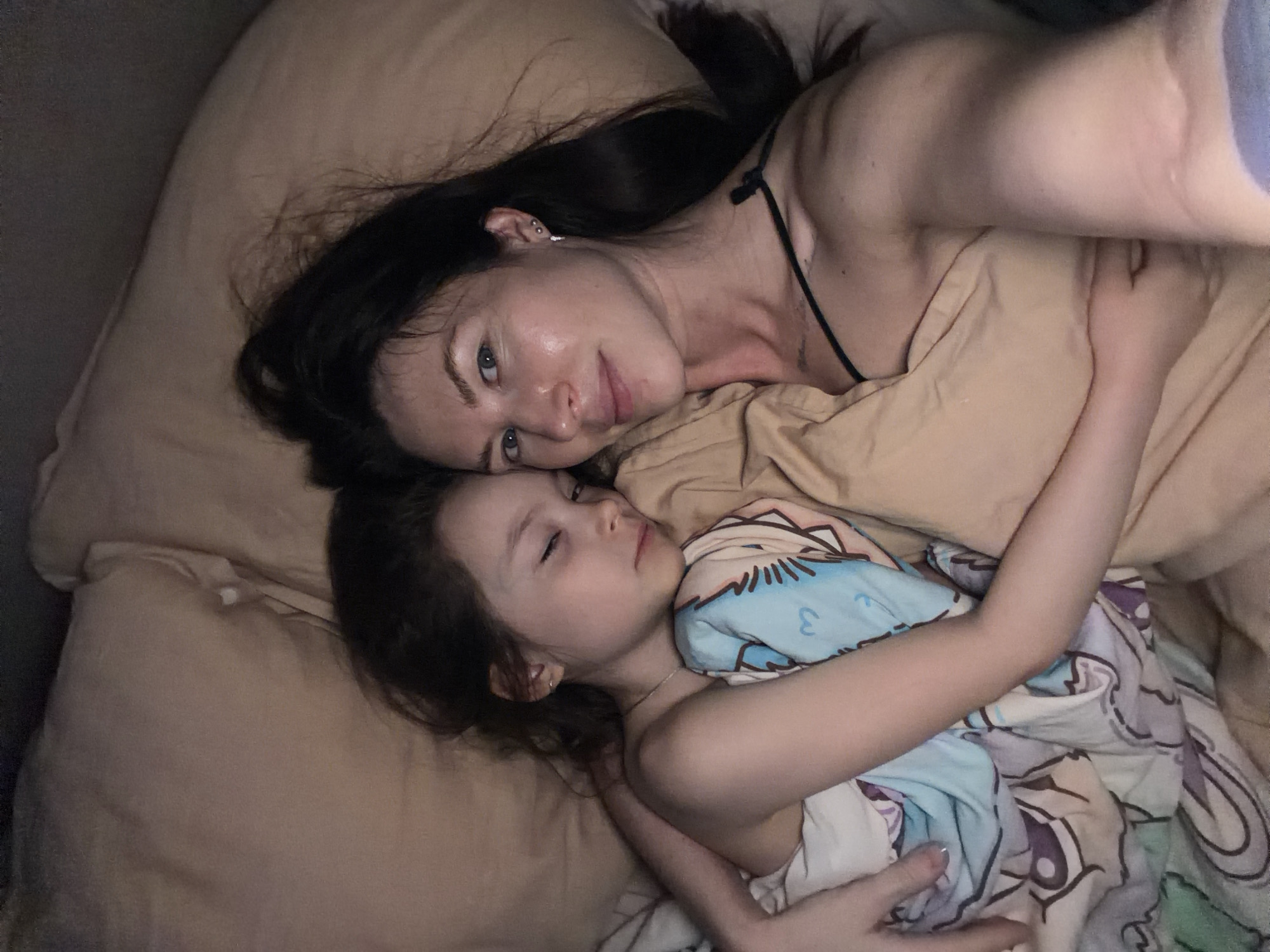 A woman and a child are lying in bed. The woman has long dark hair and is wearing a thin-strapped top. The child is under a patterned blanket and has eyes closed. The bed has beige-colored sheets and pillows. The location is Perm, Russia.