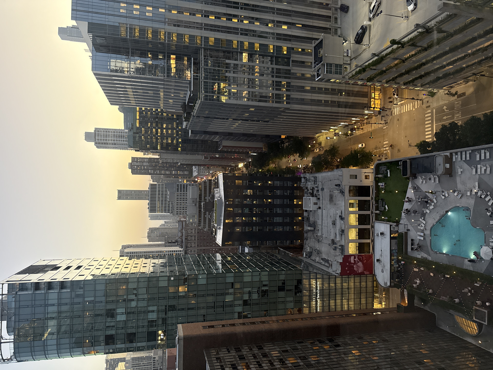 This is an aerial, eye-level, vertical, full shot of a city skyline in Chicago, Illinois, United States of America, taken at dusk. The sky is a gradient of pale orange and yellow, indicating the sun is setting. The foreground is dominated by the side of a tall, modern building with a dark, brick-like facade. To the right, a large, curved glass building reflects the twilight sky and surrounding city lights.
Below these buildings, a street is visible with traffic, streetlights, and pedestrian crosswalks. There are several other tall buildings of varying architectural styles, some with illuminated windows, extending into the distance. A multi-level parking garage with cars parked on its levels is on the right side of the image.
Further down, a rooftop amenity space is visible. This area includes a swimming pool with people swimming, lounge chairs around the pool, a grassy area with seating, and a walkway lined with string lights. People are seen scattered around this outdoor space. A large red mural of a man's face is visible on the side of one of the buildings. The overall scene depicts a bustling urban environment as day transitions into night.