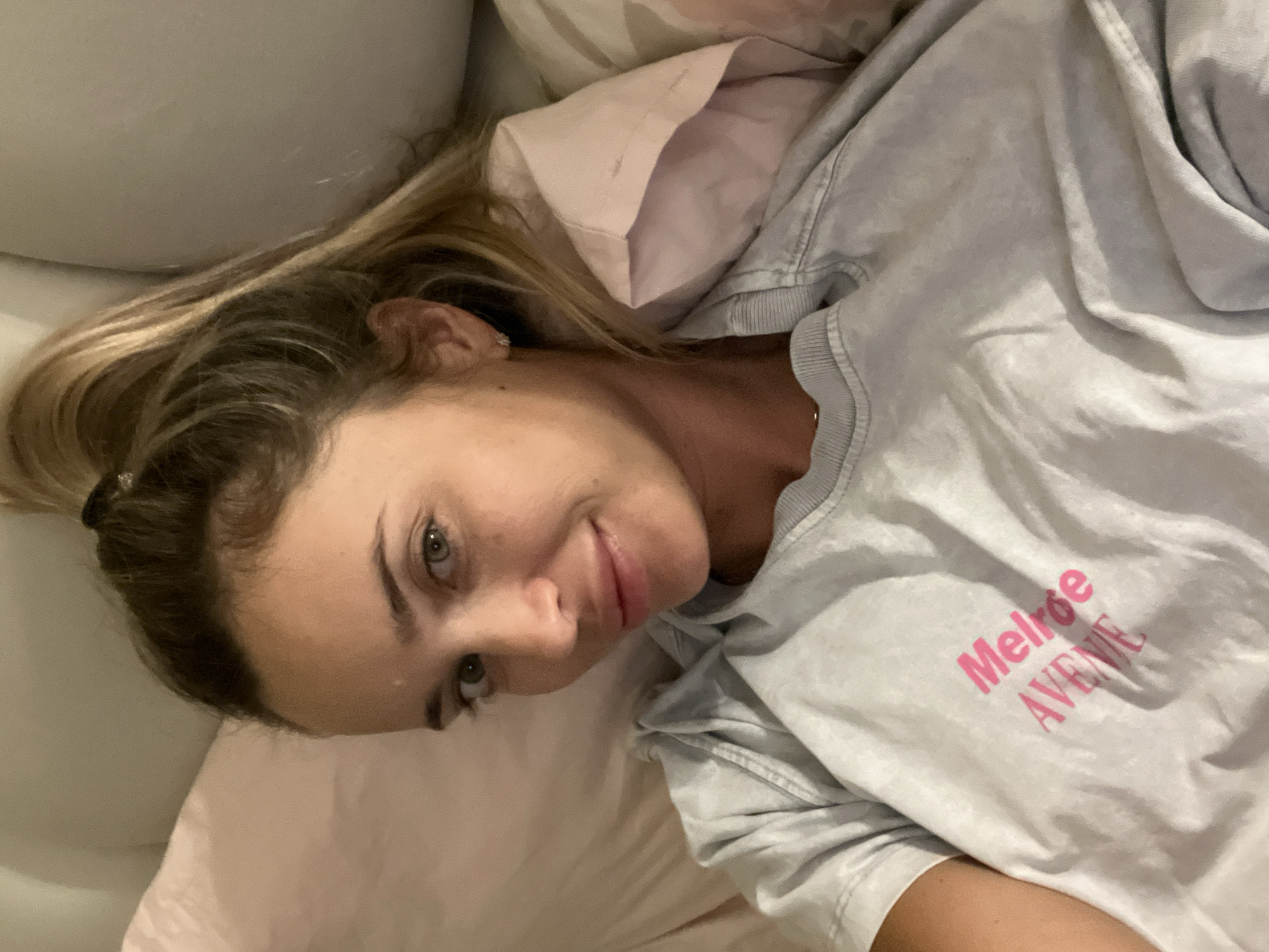 A woman with blonde hair in a ponytail is lying down and looking at the camera. She is wearing a light gray t-shirt with pink text that reads "Melroe AVENUE". The woman has green eyes and is smiling. She is in bed, with light pink pillows visible behind her. The image appears to be a selfie taken in a bedroom. There is no indication of the location being Sharm El Sheikh, Egypt.