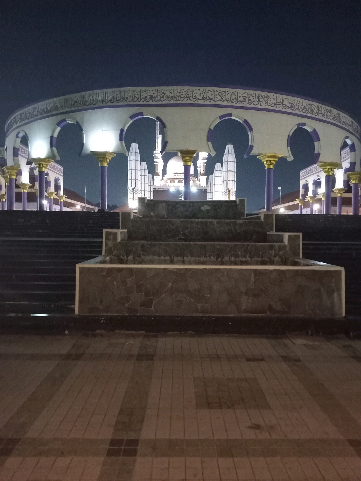 This is a nighttime photograph of the Grand Mosque of Central Java (Masjid Agung Jawa Tengah) in Semarang, Indonesia. The scene is illuminated by artificial lights, creating a dramatic and serene atmosphere.
The main subject is the mosque's grand facade, characterized by a wide, curved structure with large arches. These arches are adorned with intricate Arabic calligraphy along the top, written in white against a dark background. Supporting the curved structure are rows of tall, slender columns with purple shafts and ornate gold-colored capitals. White lights emanate from within the arches, casting a bright glow.
In the foreground, a series of dark, tiered steps leads up towards the mosque. Partially visible on these steps is a stone-tiered fountain structure. The ground in front of the steps is paved with rectangular tiles arranged in a patterned walkway.
The time of day is clearly night, with the dark sky providing a stark contrast to the illuminated architecture. There are no people visible in the image, suggesting a quiet and tranquil moment. The weather appears calm. The presence of the distinctive architecture, including the minarets visible in the background, firmly places the location as the Grand Mosque of Central Java. The overall mood is peaceful and awe-inspiring, highlighting the beauty of the mosque at night.