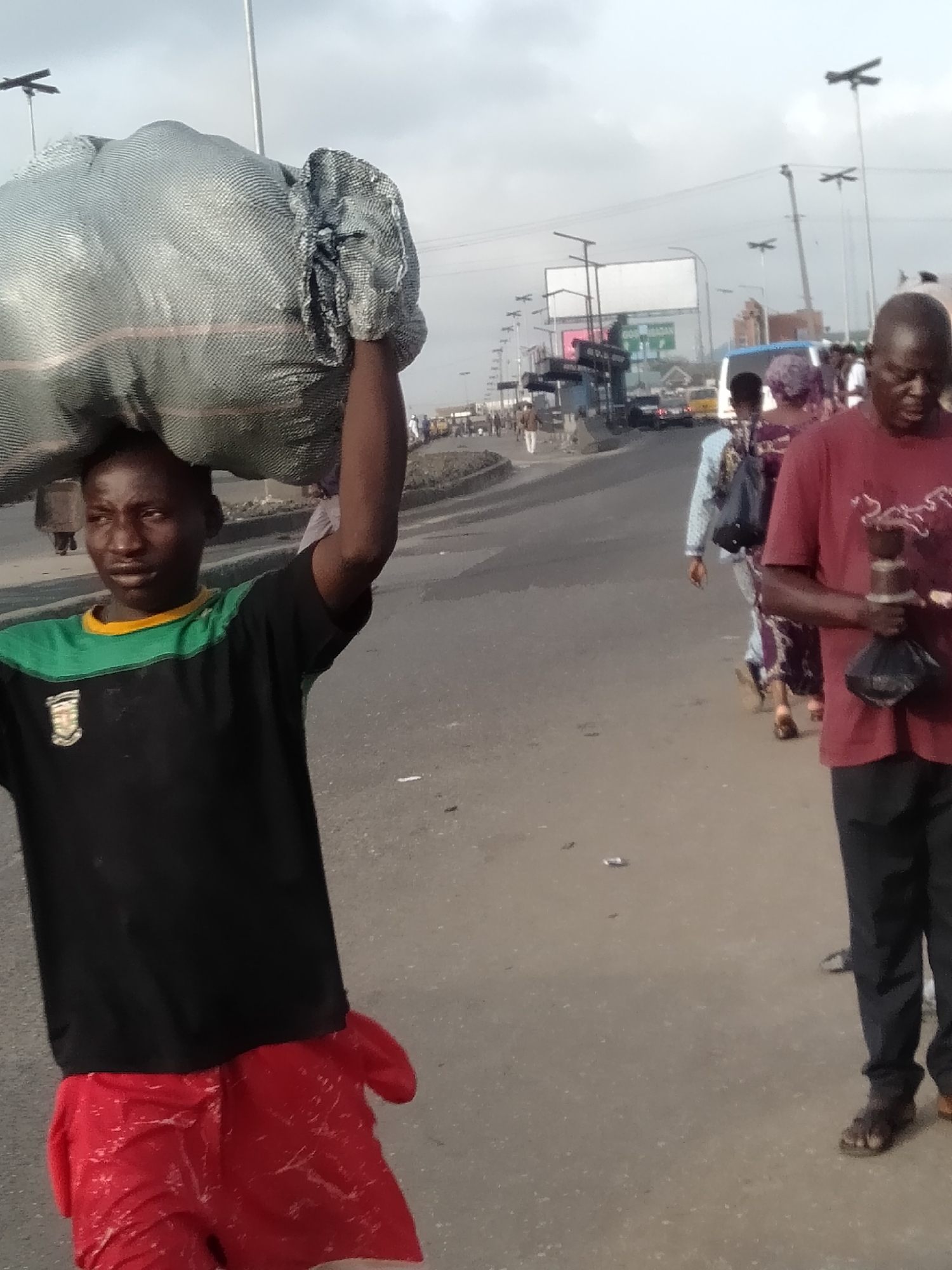 The image captures a bustling street scene in Shangisha, Nigeria, under an overcast sky during daylight hours. The primary subject, a young man, dominates the left foreground, carrying a large, light grey, woven sack on his head. He wears a black short-sleeved shirt with a green upper panel and a yellow collar, featuring an unidentifiable emblem on the left chest. His lower body is clad in bright red shorts or a wrapped cloth. His gaze is directed slightly to his right, forward.
To his right, further back, another man is visible from the waist up, wearing a red short-sleeved shirt and dark trousers. He holds a dark, possibly metallic or ceramic, object in his right hand, which appears to have a small dark bag attached or held underneath. He is looking downwards.
In the mid-ground and background, several other people are walking along the paved area beside a multi-lane road. Some are blurred, suggesting motion. One person in the mid-ground, with their back to the camera, wears a patterned outfit. Vehicles, including a light blue bus, are visible on the road in the distance. The setting features multiple utility poles or streetlights with distinctive horizontal crossbar structures extending across the road. A large, unreadable billboard stands in the far background. Small pieces of litter are scattered on the grey pavement. The overall activity suggests daily life and movement in an urban or semi-urban environment, with individuals transporting goods or commuting.