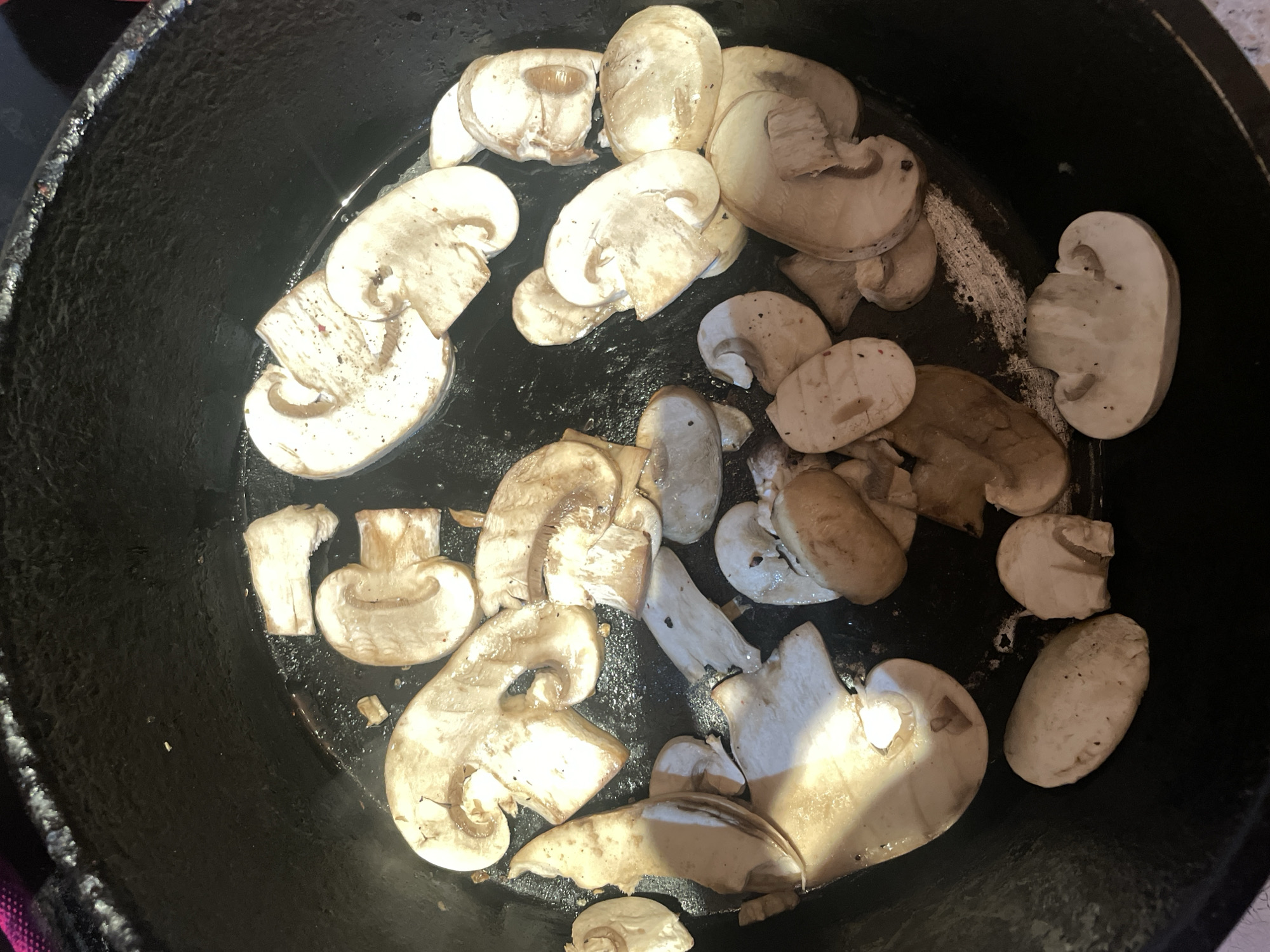 An overhead, close-up shot captures a collection of sliced mushrooms being cooked or prepared in a dark, round pan, likely a cast iron skillet. The pan's surface is black, textured, and shows some areas of dried white residue, possibly from previous cooking or cleaning.
The primary subjects are numerous white button mushrooms (Agaricus bisporus), cut into various slices and pieces, scattered across the bottom of the pan. Some slices are oval, while others are more irregular, revealing the internal structure and gills. A few mushrooms exhibit a slightly browned or translucent appearance, suggesting they are in the process of being cooked, possibly sautéed, with some liquid or oil visible reflecting light. Dark specks, potentially seasoning like pepper, are visible on some mushroom surfaces.
The lighting appears to be bright, casting some reflections on the wet surfaces of the mushrooms and the pan. There are no people visible, and no discernible text is present in the image. The scene is consistent with food preparation in a kitchen environment. While the location context provided is Novoe Devyatkino, Russia, there are no specific visual cues within the image itself to confirm this geographical location. The focus is entirely on the culinary process.