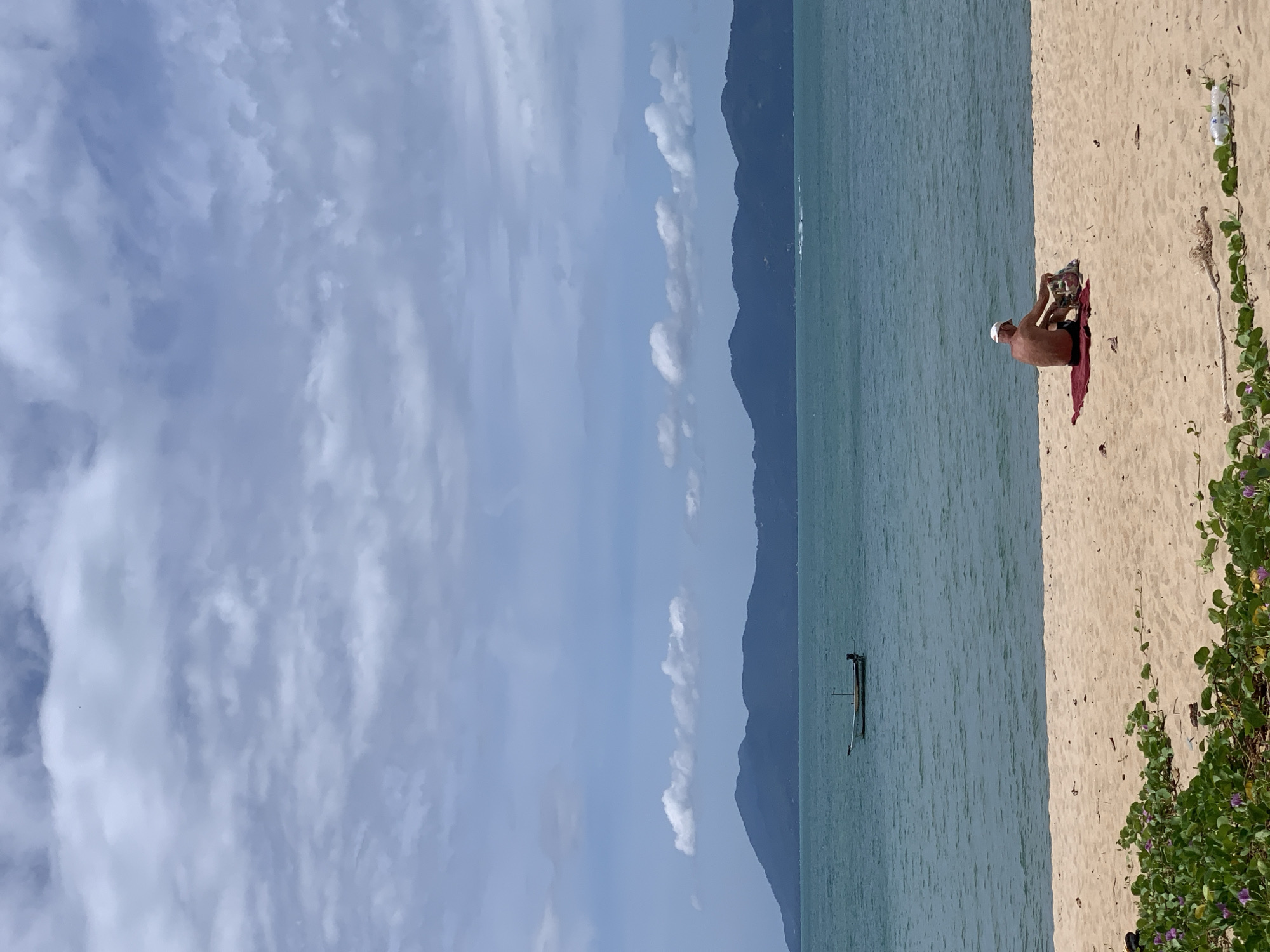 A man wearing a white cap and black shorts sits on a red towel on a sandy beach. He is facing away from the camera, towards the ocean, and appears to be reaching for something in a bag next to him. The ocean is a calm, turquoise color and a small white boat with a dark stripe is anchored offshore. In the distance, a chain of dark, tree-covered mountains forms the horizon. The sky is a soft blue with scattered white clouds. Green vines with small purple flowers grow along the edge of the beach in the foreground.