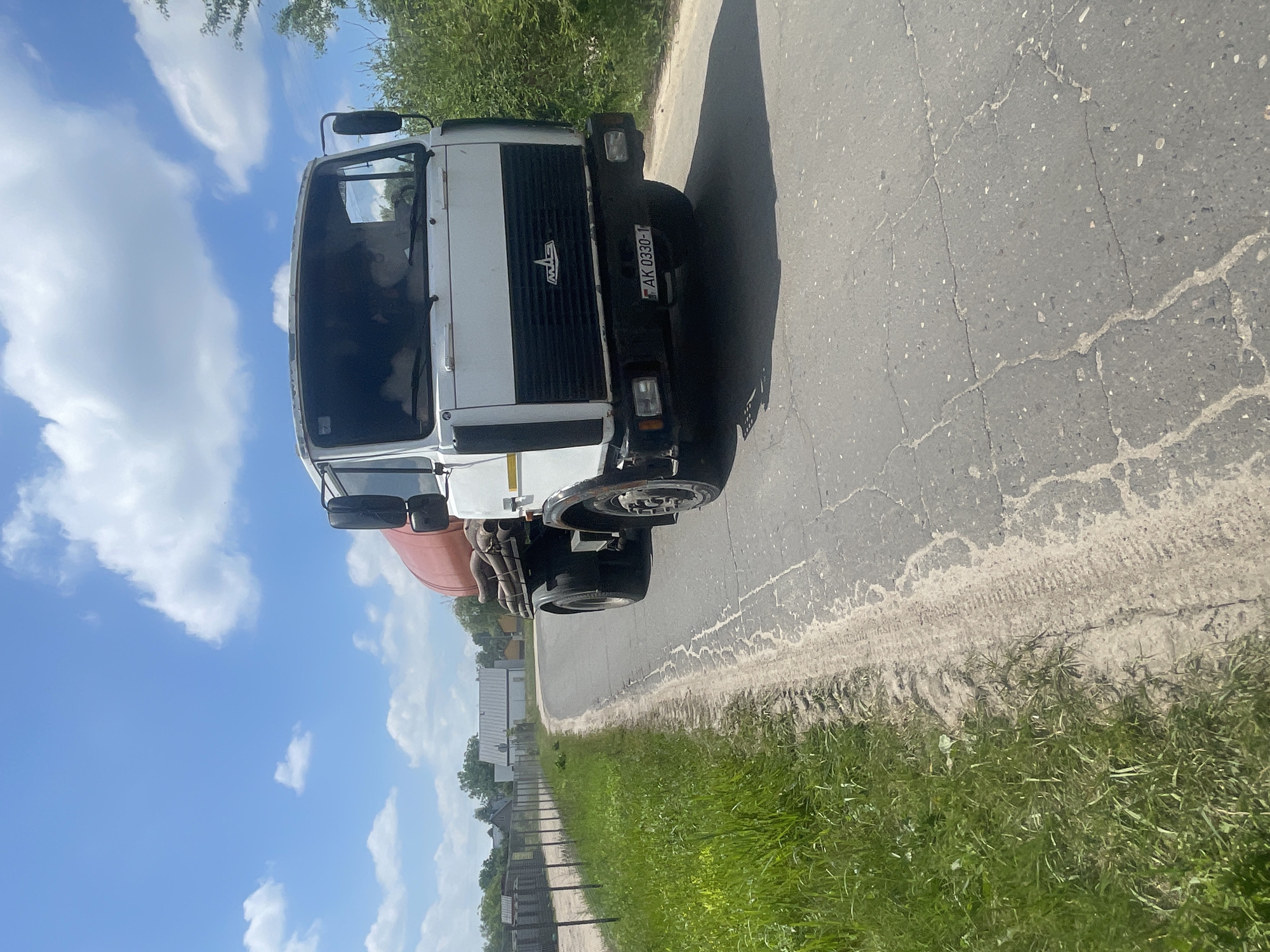 In Мухавецкі сельскі Савет, Belarus, a white MAZ truck with a red cylindrical tank is driving on a cracked asphalt road bordered by green grass and vegetation. The truck's license plate reads "AK 0330-1". Two figures are visible inside the truck's cab. In the background, there are fences, buildings, and trees under a bright blue sky with scattered white clouds.