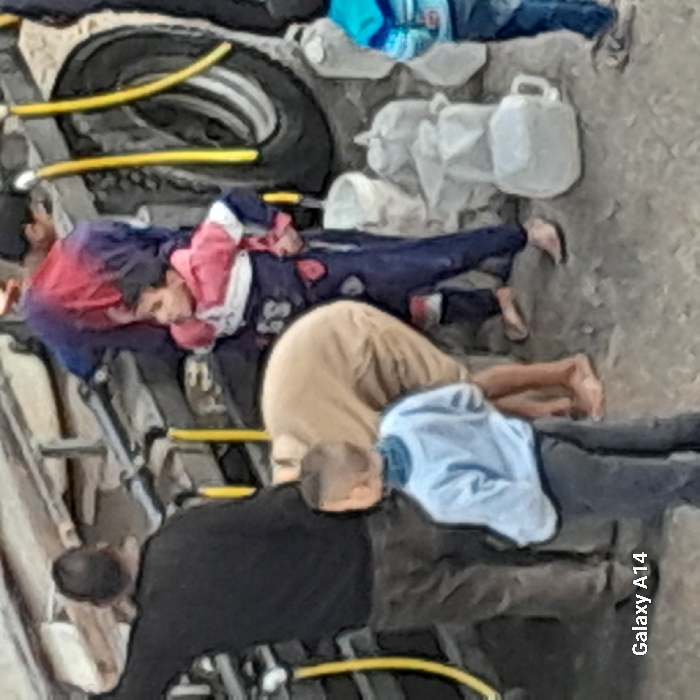 The image captures an outdoor scene in Khan Yunis, Palestinian Territory, featuring six individuals gathered around a dark-colored utility vehicle, likely a water tanker. On the far left, an adult male in a dark long-sleeved shirt and trousers is seen from behind, with his right arm extended towards the vehicle's upper structure, which includes yellow hoses. Adjacent to him, an adult in light tan attire is bent over, facing away from the camera, barefoot, interacting with something at ground level near the vehicle. In the foreground, a child wearing a light blue long-sleeved top with a darker collar and dark trousers stands with their back to the camera, hands clasped behind their back. To the right, another adult in dark blue/purple clothing is partially visible behind the vehicle. A child in a red, white, and dark blue long-sleeved top and dark blue patterned trousers stands barefoot, hands on hips, looking towards the right. A final child, partially visible on the far right, wears a blue and white striped top and blue trousers. Numerous clear or white plastic jugs, buckets, and bags, indicative of water containers, are stacked on the dirt ground near the vehicle's large black tire on the right side of the frame. Yellow hoses are prominently visible along the side of the vehicle.
