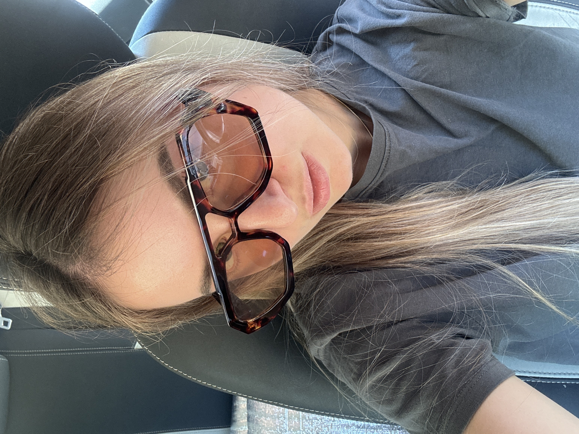 The image shows a close-up, selfie-style shot of a woman wearing sunglasses with tortoiseshell frames and a dark grey t-shirt. She is seated in what appears to be a car, with a black leather seat visible in the background. The woman has long, blonde hair with highlights. Her lips are slightly parted, and she is looking down and to the side. The lighting suggests it is daytime. There is no visible information in the image to indicate a specific location within Saint-Claude, France.