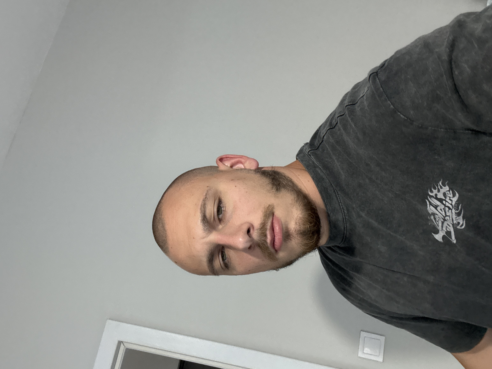 A close-up, head-and-shoulders selfie shows a man with a shaved head, light brown stubble beard, and a dark grey t-shirt with a graphic on the front. The man is looking at the camera with his head slightly tilted to his left. The background is a plain, light grey wall, with a white door frame visible to the left of the man's head. A white light switch is mounted on the wall to the left of the frame. The image appears to have been taken indoors, possibly in a residential setting in Stara Zagora, Bulgaria.
