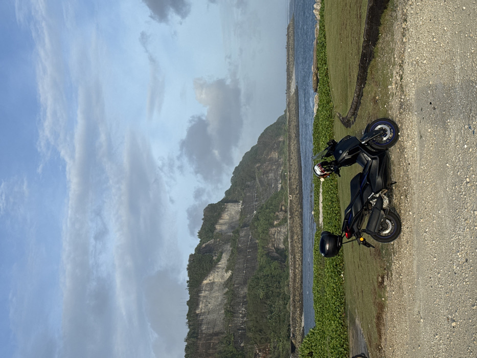 A dark blue Yamaha XMAX 300 maxi-scooter, fitted with a black top case and a white, red, and black full-face helmet resting on its front, is parked on a light-colored gravel surface. The scooter's front wheel features a blue rim accent. To its right and behind it, a manicured green lawn is visible, bordered by a low, dark stone or concrete barrier. Beyond the lawn, the deep blue ocean is observed with active waves. A long, dark grey rock breakwater extends into the sea from the left, separating calmer coastal waters. In the midground, a strip of dense green foliage lines the shoreline. The background is dominated by towering, stratified grey cliffs, extensively covered with green vegetation, particularly on their upper sections. A narrow path or road is visible traversing the face of the cliffs. The sky is light blue with scattered white and grey clouds. No individuals are present in the scene, which is located in Ungasan, Indonesia.