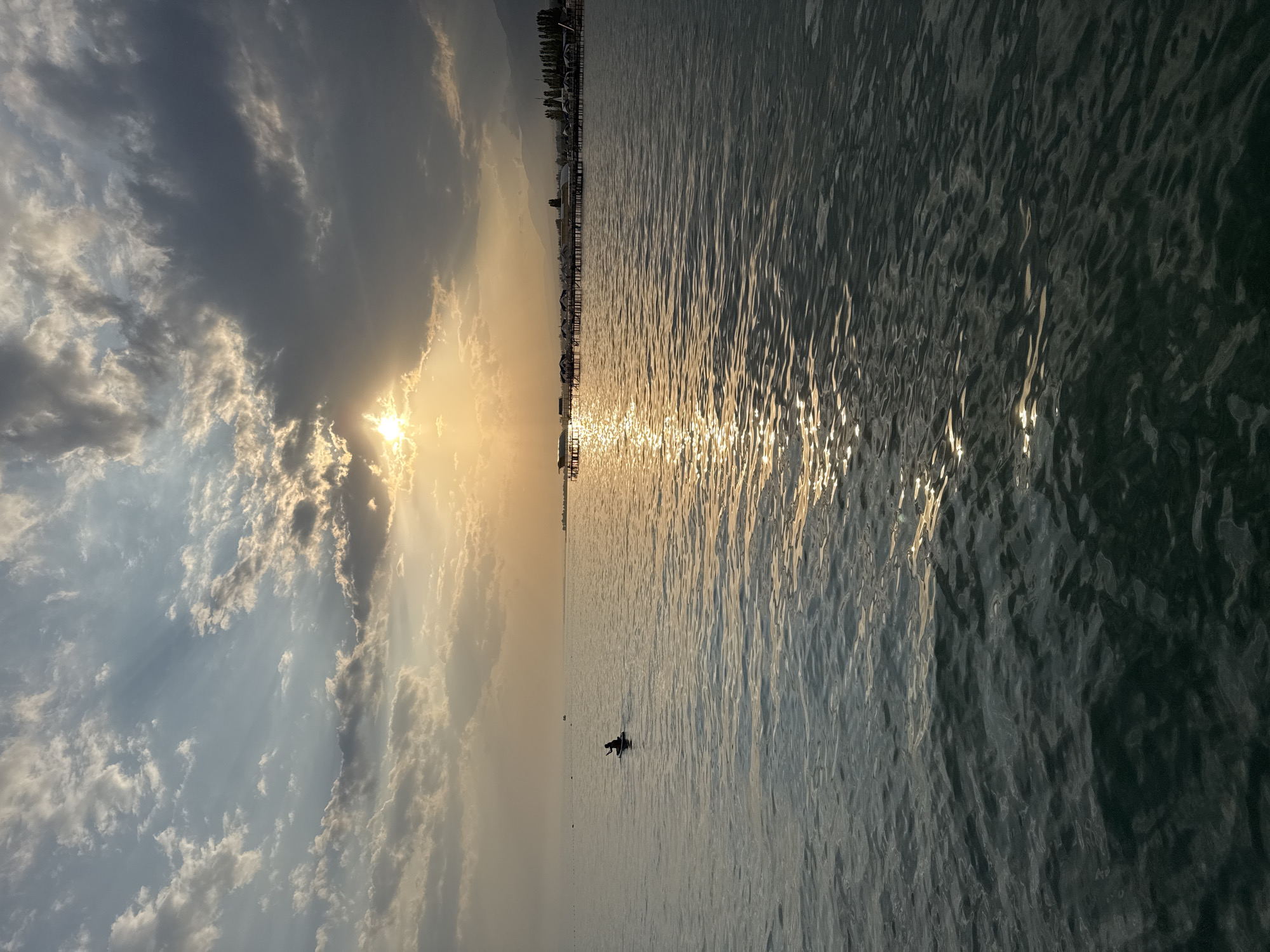 A tranquil scene unfolds at sunset over Issyk-Kul Lake in Sary-Oy, Kyrgyzstan. The sky is a dramatic canvas of clouds, with the sun breaking through, casting golden rays across the water. In the foreground, the rippling surface of the lake reflects the warm hues of the setting sun.
Further out, a lone figure is silhouetted on a paddleboard, peacefully gliding across the calm water. The distant shoreline is dotted with structures, including a long pier extending over the lake, lined with small buildings or cabanas, and a cluster of trees. Mountains form a hazy backdrop against the horizon, adding to the serene landscape. The overall impression is one of quiet solitude and the beauty of nature at the end of the day.
