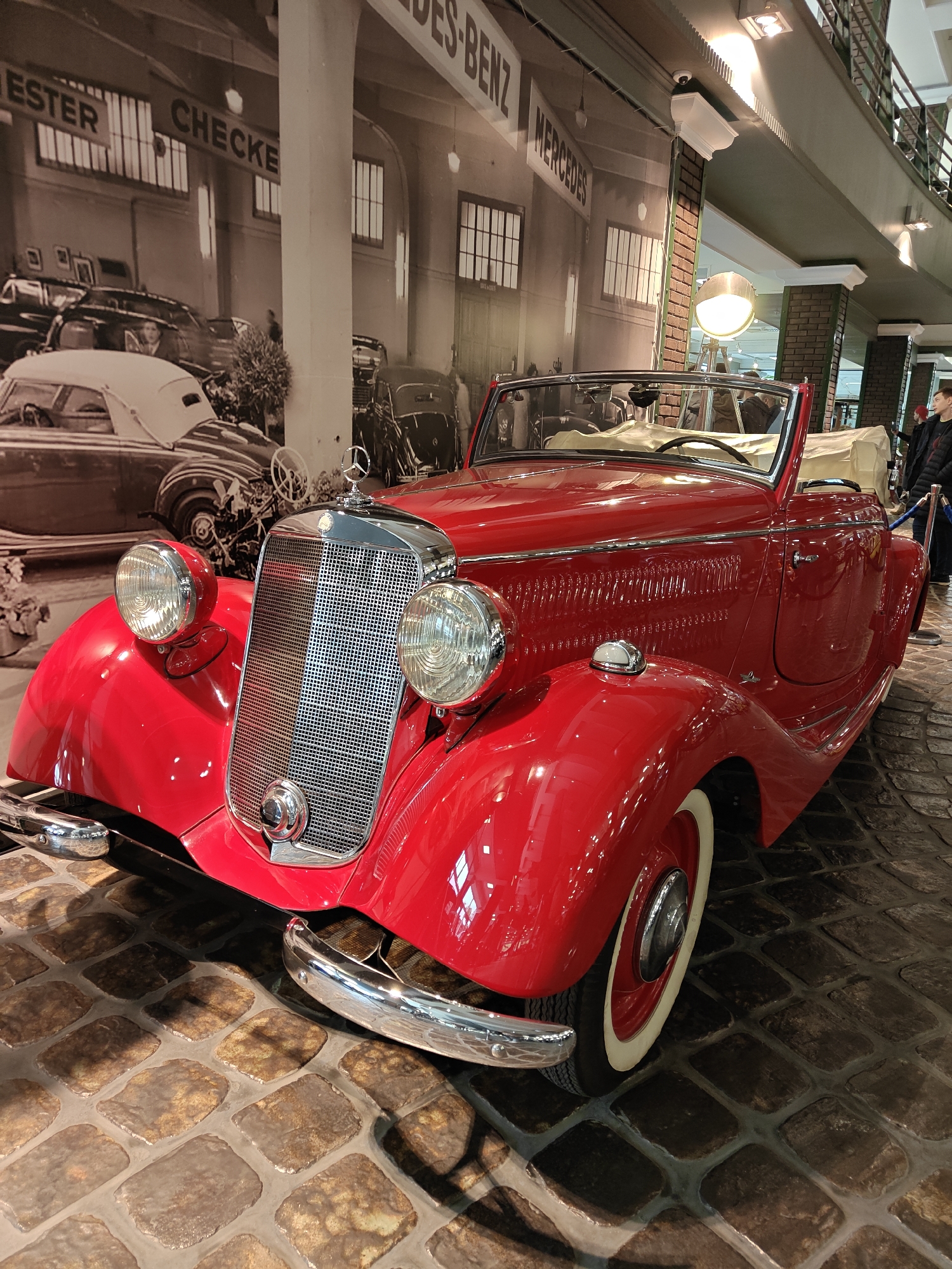 The image shows a red vintage Mercedes-Benz convertible displayed on a cobblestone floor. The car has a polished chrome grille and whitewall tires. Behind the car is a black and white mural depicting a Mercedes-Benz dealership or garage, with "ESTER CHECKER" and "MERCEDES-BENZ" visible on the building. A large industrial-style lamp is positioned near the car. A person is standing in the background, near a blue rope or barrier. The scene appears to be inside a museum or exhibition space. The location mentioned, Архангельское, Russia, might be where the car is displayed, although not directly visible in the image.