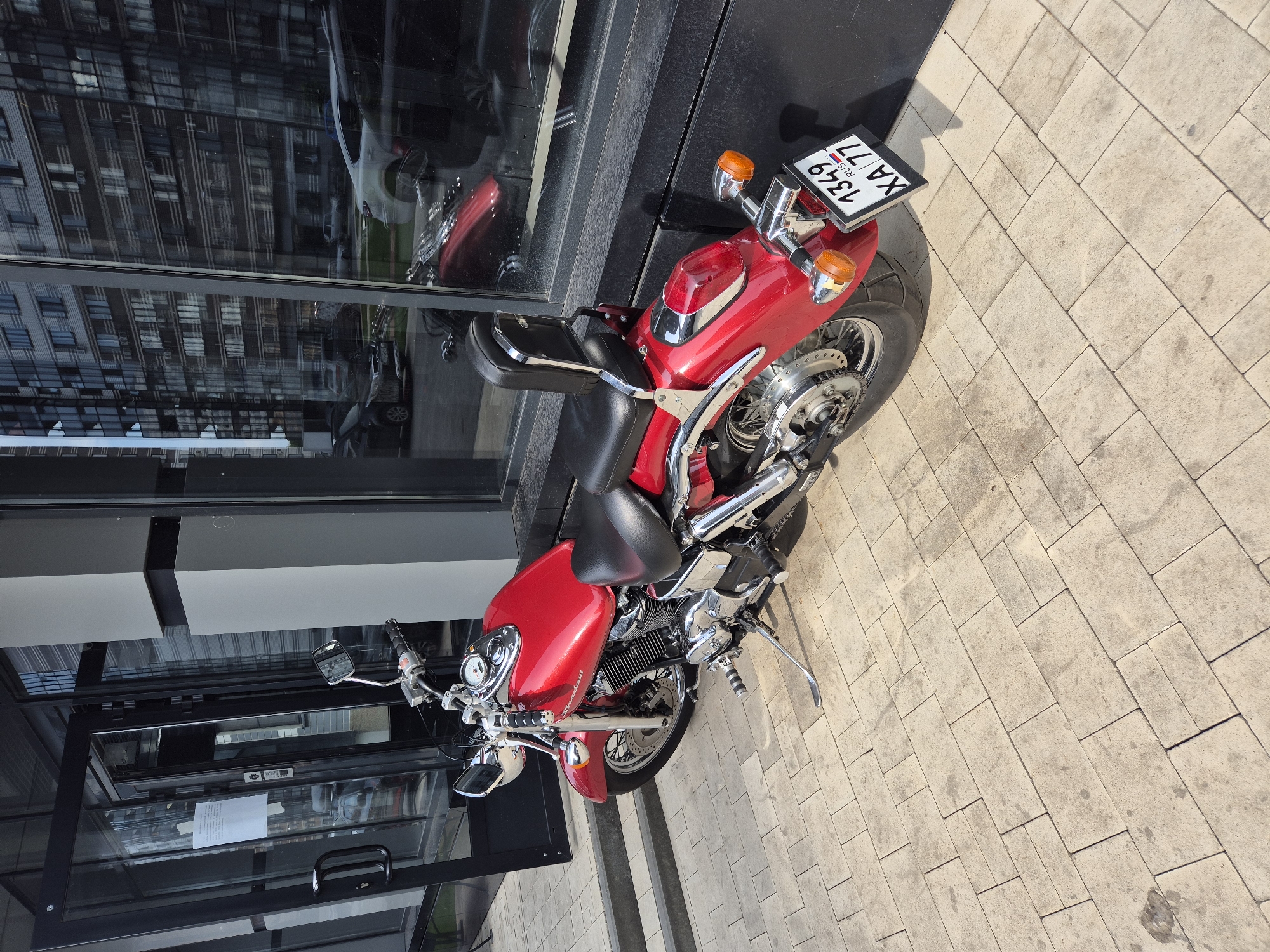 A red motorcycle is parked on a paved walkway outside a modern building with a glass facade. The motorcycle is a cruiser style with a black seat and a passenger backrest. A license plate with the characters "1349 XA 77" is visible on the rear of the motorcycle. The building's windows reflect apartment buildings, suggesting an urban setting. The walkway is made of light-colored rectangular pavers. The scene appears to be in the city of Voronezh, Russia, as indicated by the license plate format which is characteristic of Russian plates.