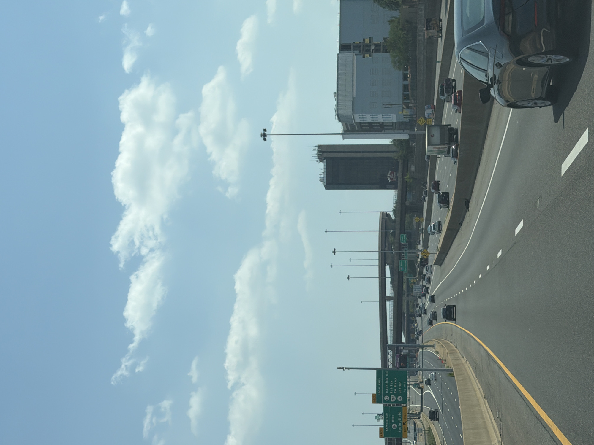 The image shows a view from a moving vehicle on a multi-lane highway, likely in Springfield, United States of America, as indicated by road signs. The sky is blue with scattered white clouds. The highway has concrete barriers separating lanes and the direction of travel. Several vehicles, including cars and a truck, are visible on the highway. Overhead signs indicate exits for "Springfield," "Express Exit," "Local Exits," "Backlick Rd," "Fairfax," "Co Pkwy," and "Lorton." In the background, several buildings of varying heights, including what appear to be office buildings, are visible, along with lampposts and overpasses. The environment appears to be urban or suburban.
