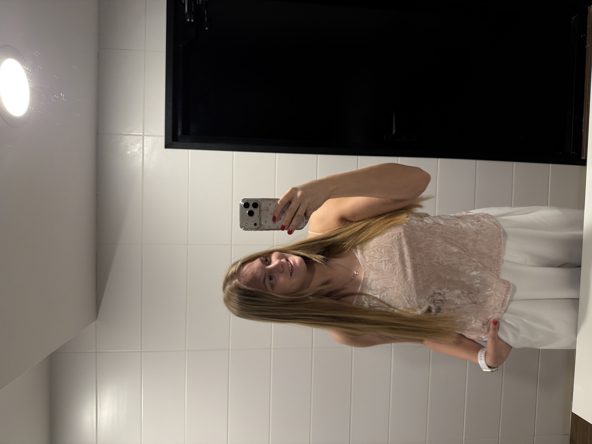 A woman is taking a selfie in what appears to be a bathroom. The background consists of white tiles and a black cabinet above the sink. The woman is holding a phone in her right hand. She has long, blonde hair and is wearing a light pink top and white pants.
Location: No evidence to support it being in Izhevsk, Russia.