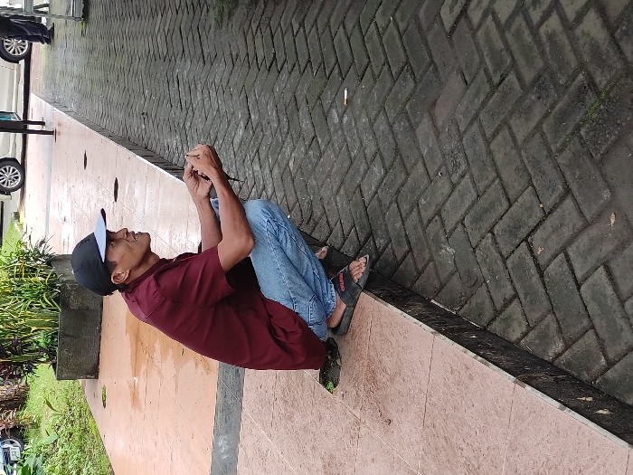 A man is depicted in a crouching or squatting position outdoors, possibly in Kepanjen, Indonesia. He wears a dark red short-sleeved collared shirt, light blue denim jeans, black flip-flops, and a black baseball cap with a light blue brim. He is looking upwards with a focused expression, holding a small, dark object in both hands, which he appears to be examining or tending to.

The setting is a paved area, possibly a sidewalk or pathway. The ground beneath the man consists of light brown tiles or a similar smooth surface. Adjacent to this, separated by a low curb or edge, is a section of darker grey interlocking pavers laid in a herringbone pattern. Some patches on the ground appear damp. In the background to the left, green foliage from bushes or plants is visible, along with parts of parked vehicles, including a white car and a dark-colored car.

The time of day is daytime, with soft, diffused lighting suggesting an overcast sky. A discarded cigarette butt can be seen on the darker paver section of the ground. The brand "CROCS" might be faintly visible on the strap of the man's flip-flops.