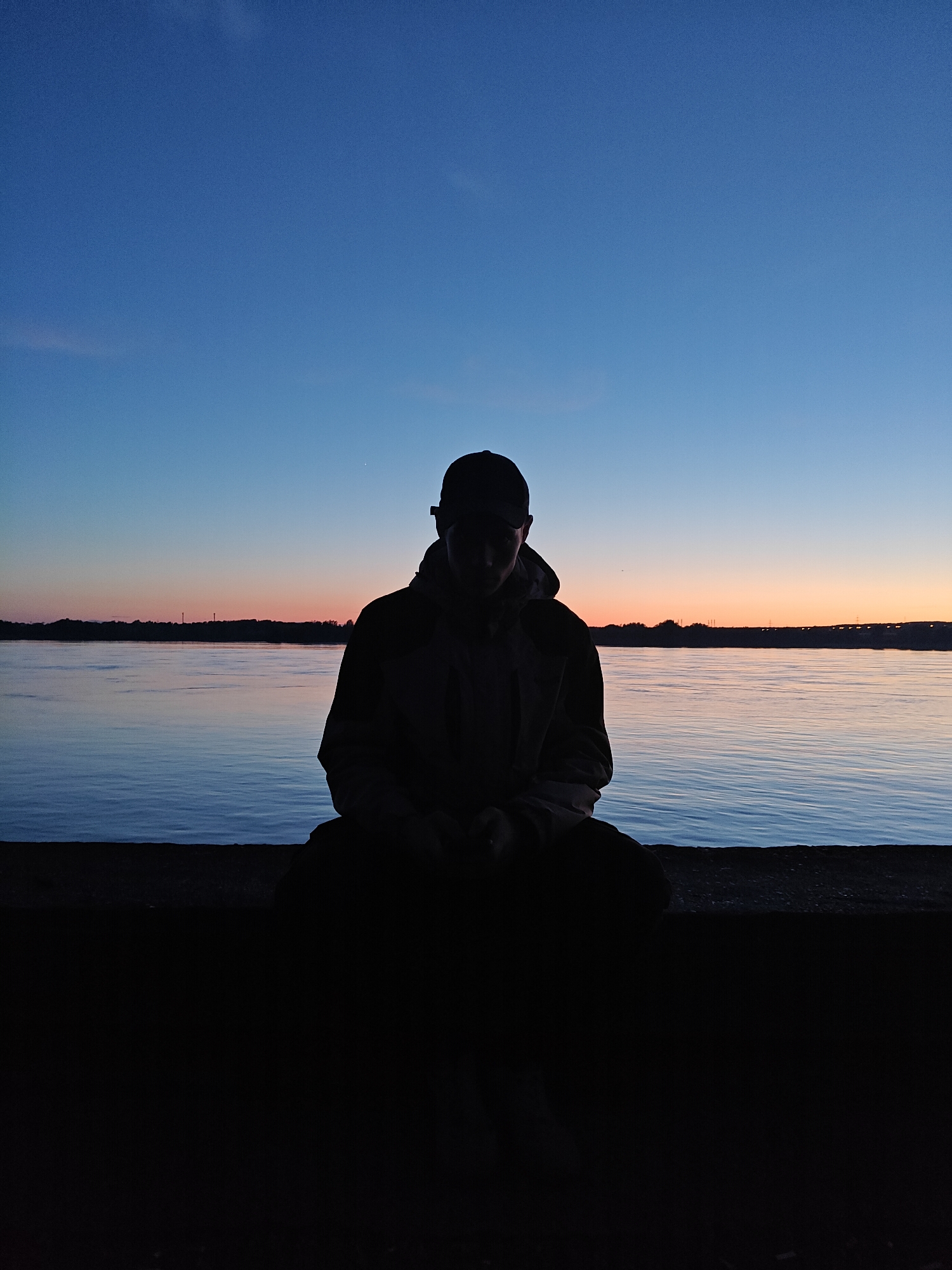 The image captures a solitary figure silhouetted against a serene twilight landscape in Chaikovsky, Russia. In the foreground, a person, likely male, is seated on a low, dark structure, possibly a wall or embankment. The individual is wearing a baseball cap and a hooded jacket, with their entire form rendered as a dark silhouette against the brighter background, indicating the light source is behind them. Their posture appears relaxed and contemplative, with hands resting in front.
The upper portion of the image is dominated by a clear sky, transitioning in color from a deep, rich blue at the top to lighter blues, and then soft oranges and pinks along the horizon, characteristic of sunset or early dusk. Below the horizon, a wide expanse of calm water reflects the sky's gentle colors, with subtle ripples visible on its surface. Across the water, a distant shoreline is visible, featuring a dark line of trees and a few indistinct structures, possibly industrial towers or buildings, against the glowing horizon.
The scene conveys a peaceful and quiet atmosphere, suggesting a moment of solitude or reflection by the water's edge. There is no visible text or identifiable weather patterns beyond a clear sky.