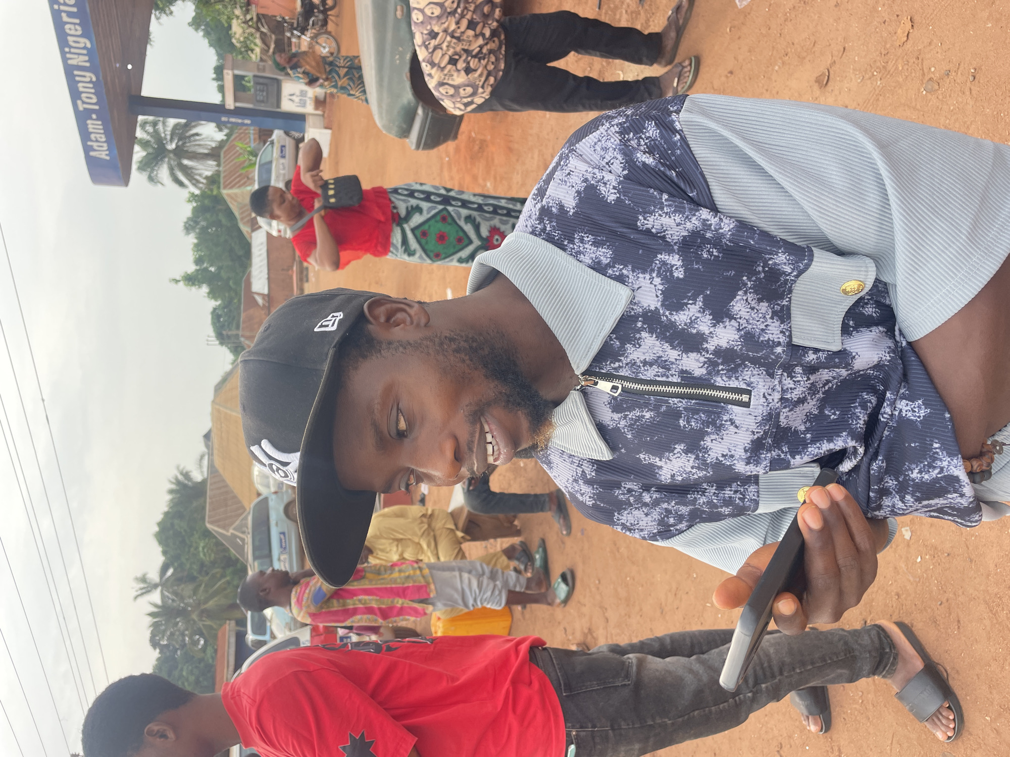 A medium-angle shot of a man in a black baseball cap and a unique button-down shirt holding a black smartphone. The man is smiling and appears to be looking at the phone. Several other individuals are visible in the background, some wearing brightly colored clothing and traditional patterns. The location seems to be a bustling area with vehicles and buildings in the distance. A sign that says "Adam-Tony Nigeria" is visible. The ground appears to be a dirt or clay surface.