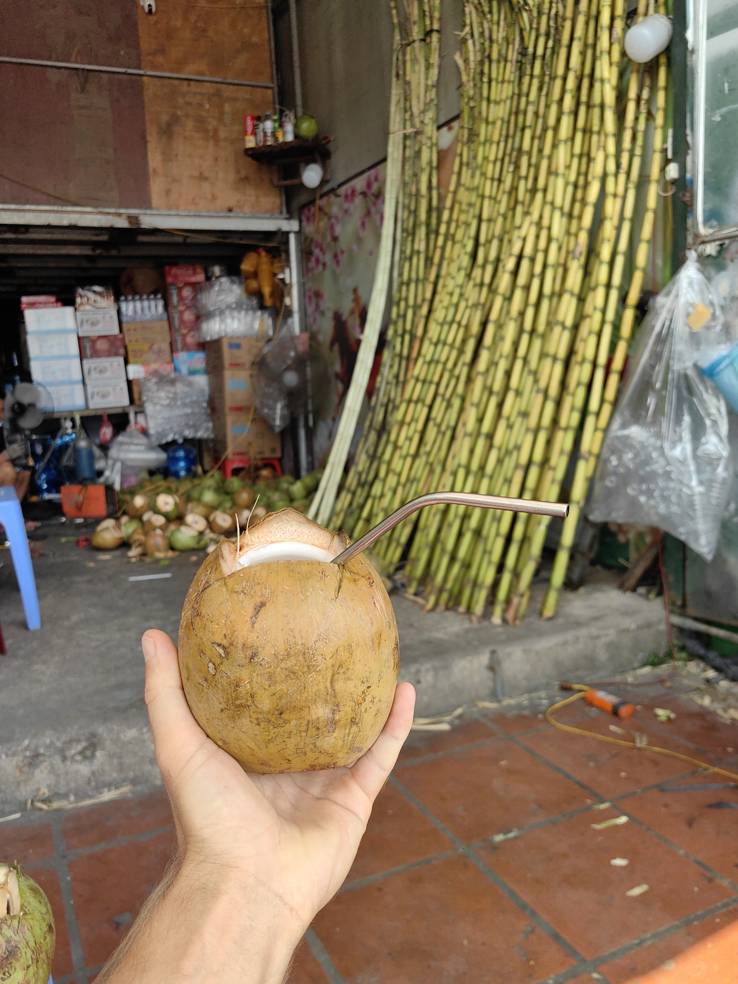 A first-person perspective shows a human hand holding a brown coconut with a metal straw inserted into the top. In the background, there are stalks of sugar cane leaning against a wall, with more coconuts piled near a storefront. The storefront appears to sell drinks and other items. The scene is located in Cao Xanh Ward, Vietnam.