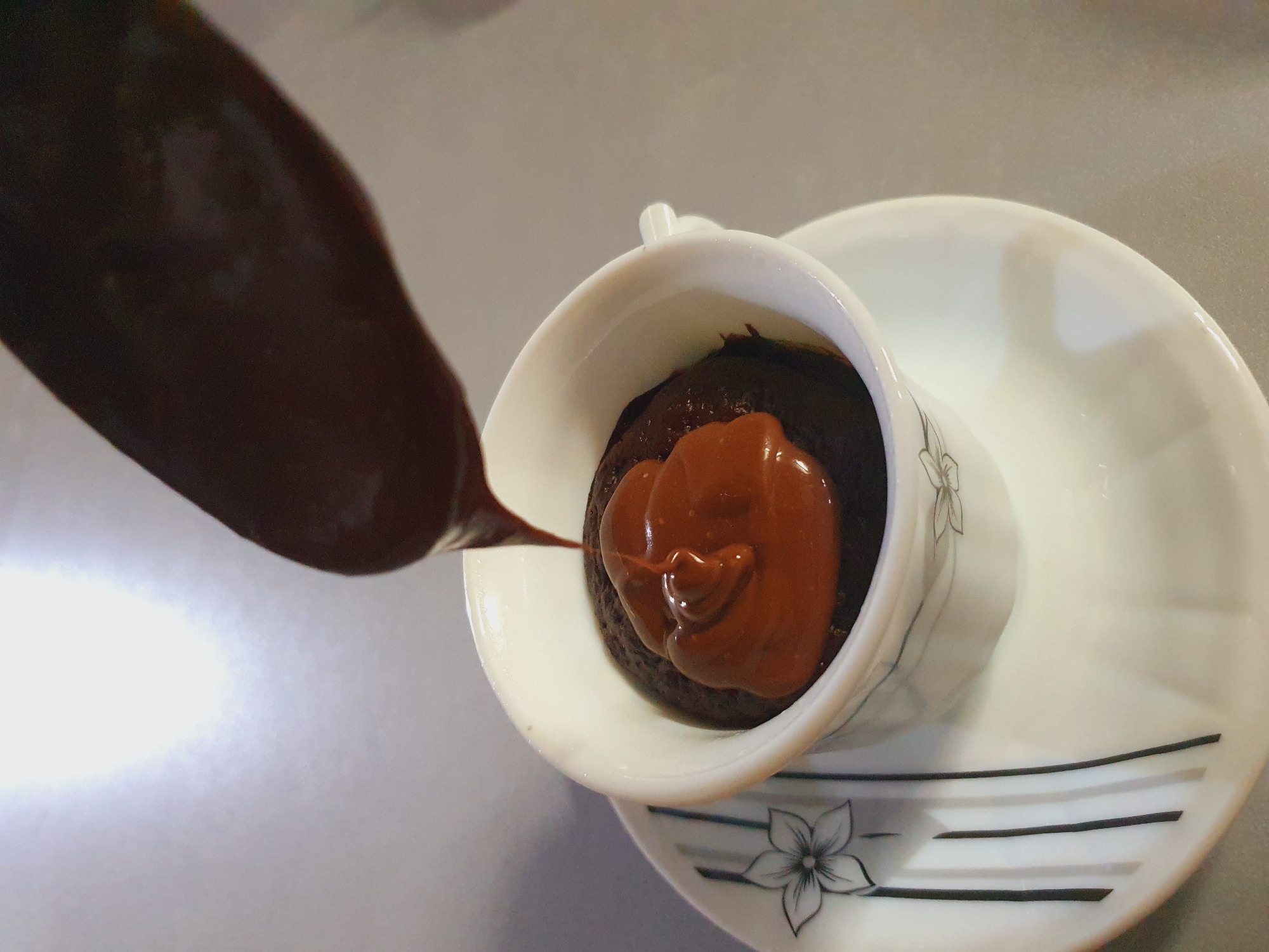 A spoon, held by an unseen hand, is drizzling melted chocolate over a dark chocolate cake baked in a white teacup. The teacup sits on a matching white saucer adorned with a floral and striped design. The background is a neutral, out-of-focus surface, likely a table. The scene depicts the preparation or serving of a dessert. There is no discernible connection to Shiraz, Iran.
