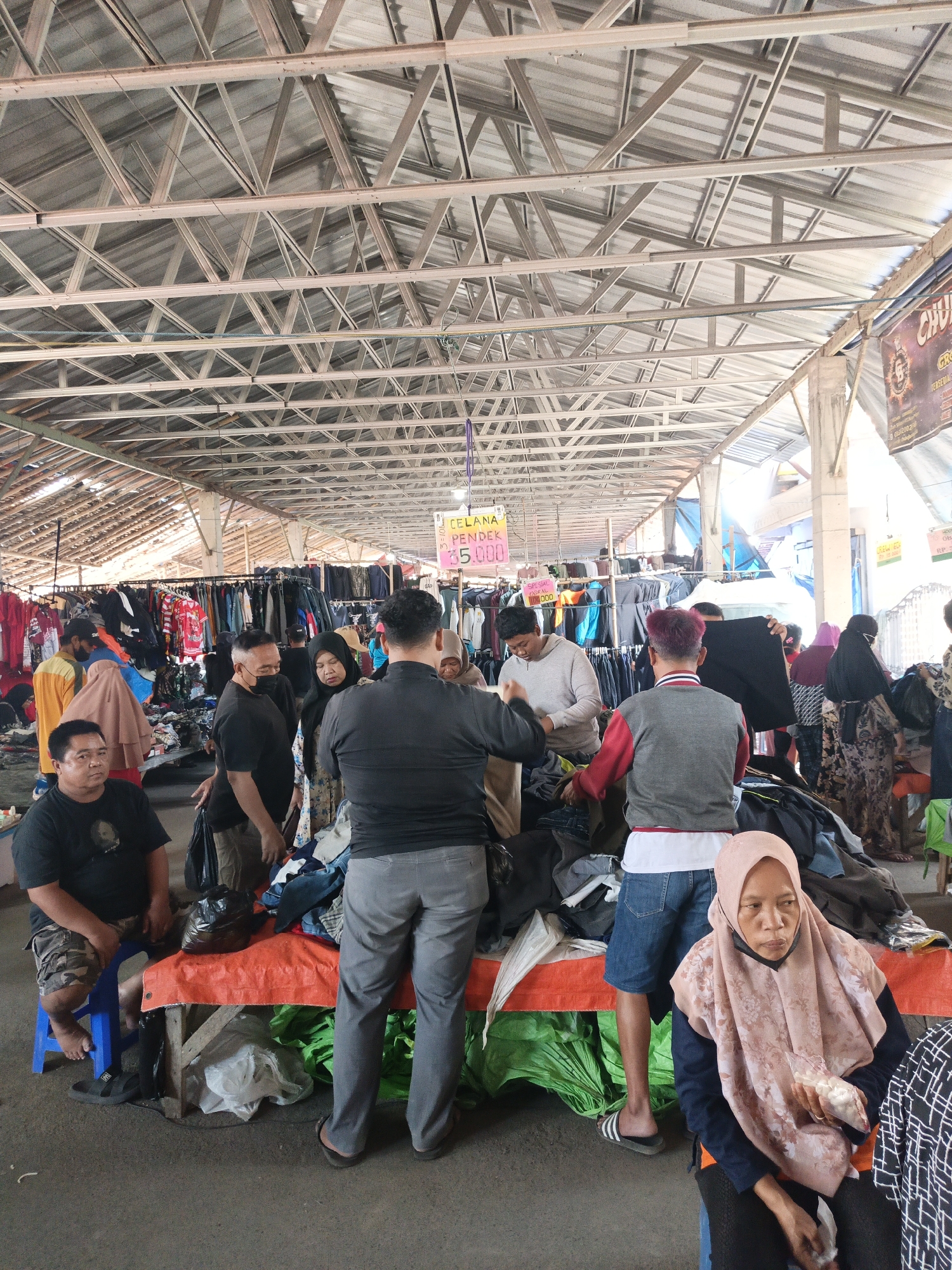 The image depicts a bustling outdoor market in Jatinom, Indonesia, likely a flea market or a local bazaar. The scene is under a large, corrugated metal roof supported by a complex metal truss system, suggesting a covered market. In the foreground, several people are engaged in shopping and selling activities.
On the left, a man wearing a black t-shirt and camouflage shorts sits on a blue stool, observing the surroundings. Next to him, another man in a black t-shirt and black mask is looking at clothes laid out on an orange tarp covering a table. A woman in a hijab, with a floral patterned dress, stands near him, also examining merchandise.
In the center of the image, a man in a grey sweatshirt is holding up a black garment, seemingly inspecting it. A man with red-dyed hair and a red and grey jacket stands behind him, facing away from the camera. Another man in a grey t-shirt and blue jeans is browsing through clothes at the table.
To the right, a woman wearing a light pink hijab and a patterned dress is seated and holding a small bag. She appears to be a vendor or a shopper. Further back, more people are visible amidst racks of clothing and various market stalls.
The market appears to be selling clothing, with numerous garments hung on racks and displayed on tables. The ground is paved, and the overall atmosphere suggests a busy commercial environment. A sign is visible in the background with text that includes "CELANA PENDEK" (short pants) and a price, indicating a retail transaction. Another sign further up reads "CHURRO". The lighting is natural, indicating it is daytime.