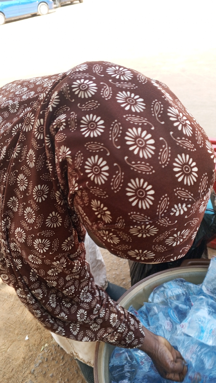 A close-up, high-angle shot captures a person leaning forward in an outdoor setting in Dakata, Nigeria. The individual wears a brown garment with a repeated pattern of white flowers and paisley shapes, covering their head and right arm. Their right hand is extended into a large, round, light-colored basin. Inside the basin, numerous small, clear plastic sachets filled with liquid, presumably water, are visible. The background shows a light-colored, possibly sandy or paved ground, and parts of two vehicles, including a blue car, are indistinctly visible in the upper left. The scene suggests a street vendor or market activity.