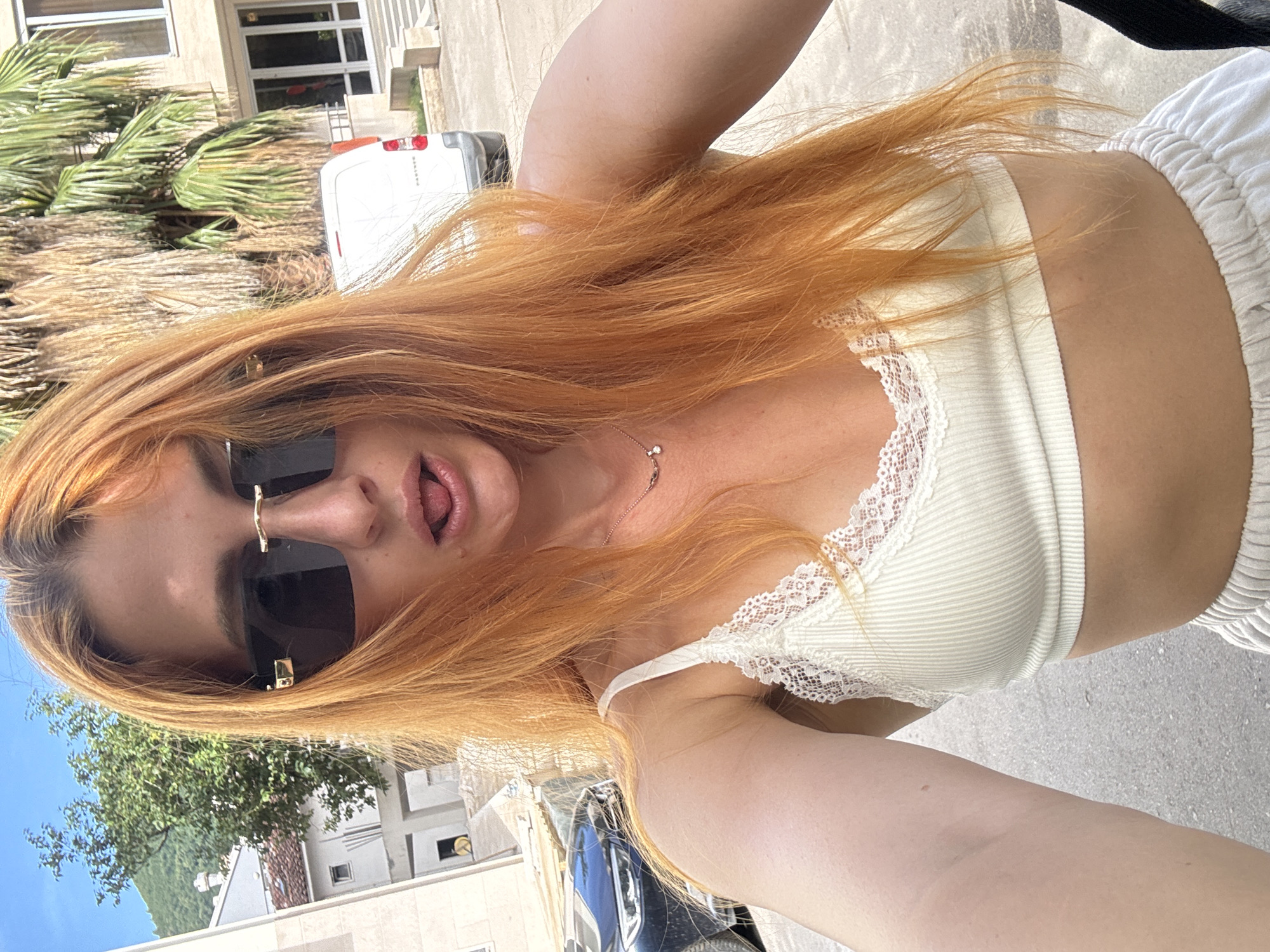 A young woman with bright ginger hair stands outdoors in Budva, Montenegro, taking a selfie. She is wearing a white lace-trimmed bralette and white drawstring shorts. Her mouth is slightly open, and her tongue is visible. She has large, dark sunglasses on, and a delicate necklace with a small pendant is visible around her neck. Behind her, a white van and a dark blue car are parked on a paved surface. Buildings and trees, including a palm tree, are visible in the background, suggesting an urban or suburban setting. The sky is blue and clear.