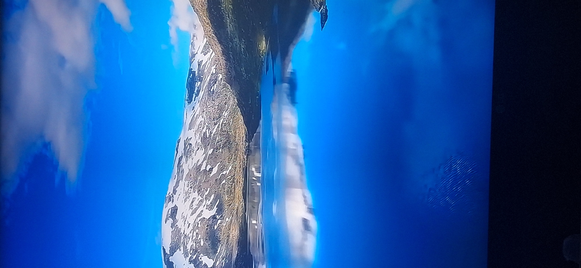 The image displays a serene natural landscape under a bright blue sky dotted with white clouds. A calm, reflective body of water dominates the lower half of the frame, mirroring the snow-capped mountains and rocky slopes that form the background. The mountains show patches of snow and rocky terrain, with some vegetation visible on the lower slopes. The water's surface is smooth, creating a clear reflection of the sky and mountains. The image does not depict the city of Bila Tserkva, Ukraine, or any urban elements.