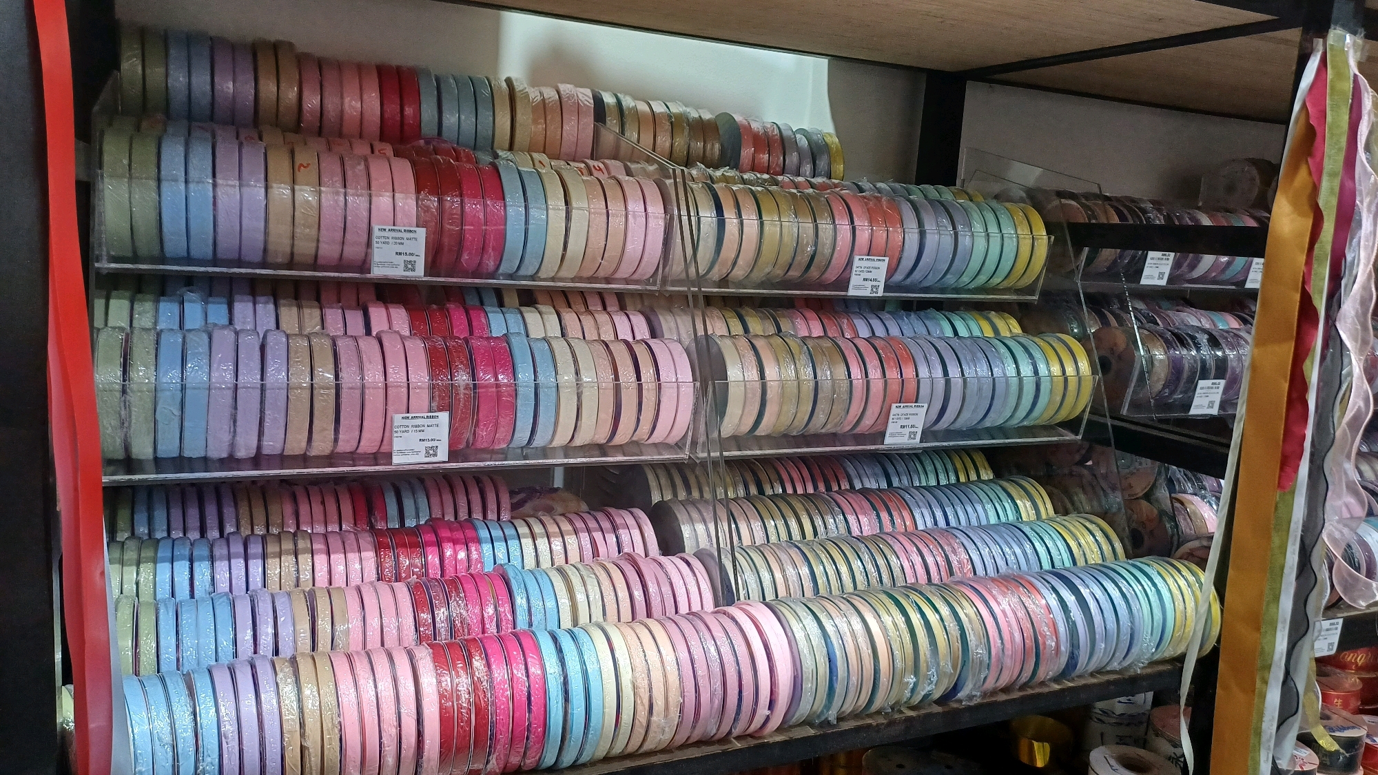 The image shows a close-up, eye-level view of shelves filled with spools of ribbon, likely in a fabric or craft store in Kuala Lumpur, Malaysia. The ribbons are organized by color and are stacked in rows on clear plastic shelves, some of which are angled. The colors range from pastel shades of pink, blue, lavender, and yellow to more vibrant reds and greens. Several price tags are visible, indicating that the ribbons are for sale. In the upper right corner, several longer, wider ribbons in various colors hang down, suggesting they are also part of the store's inventory. The overall impression is one of abundance and variety in crafting supplies.
