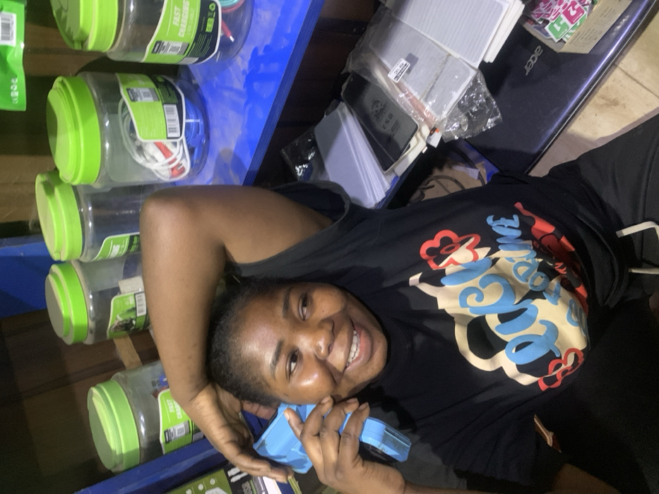 A woman smiles while holding a blue, boxy object to her ear, presumably mimicking a phone. She lies next to a blue shelf filled with plastic containers of phone chargers. An Acer laptop and other electronics are visible near her. She wears a black t-shirt with a large, colorful graphic design. The scene appears to be indoors, possibly in a shop or storage area.